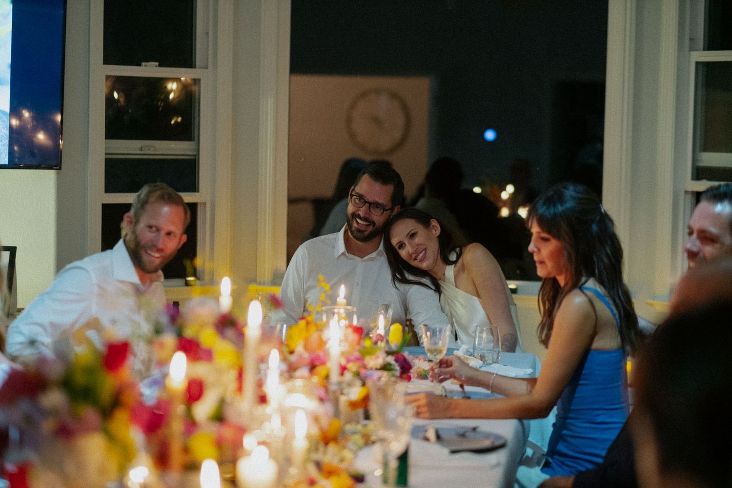 Friends laughing and celebrating together at a candlelit dinner party with colorful floral centerpieces.
