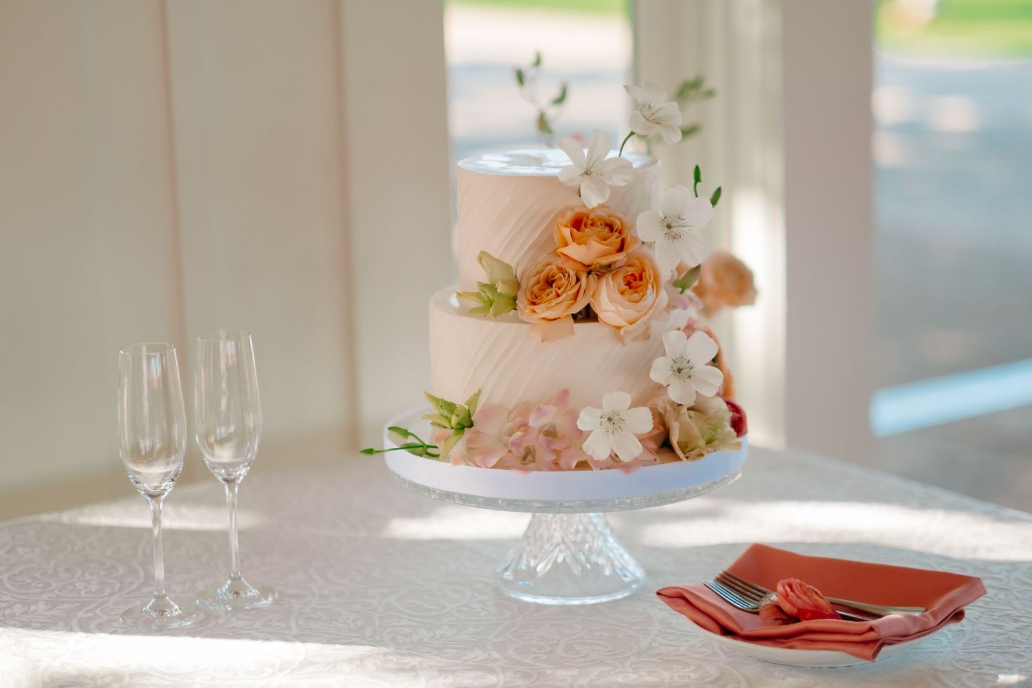 Elegant two-tier wedding cake adorned with peach roses and white flowers on a crystal cake stand.