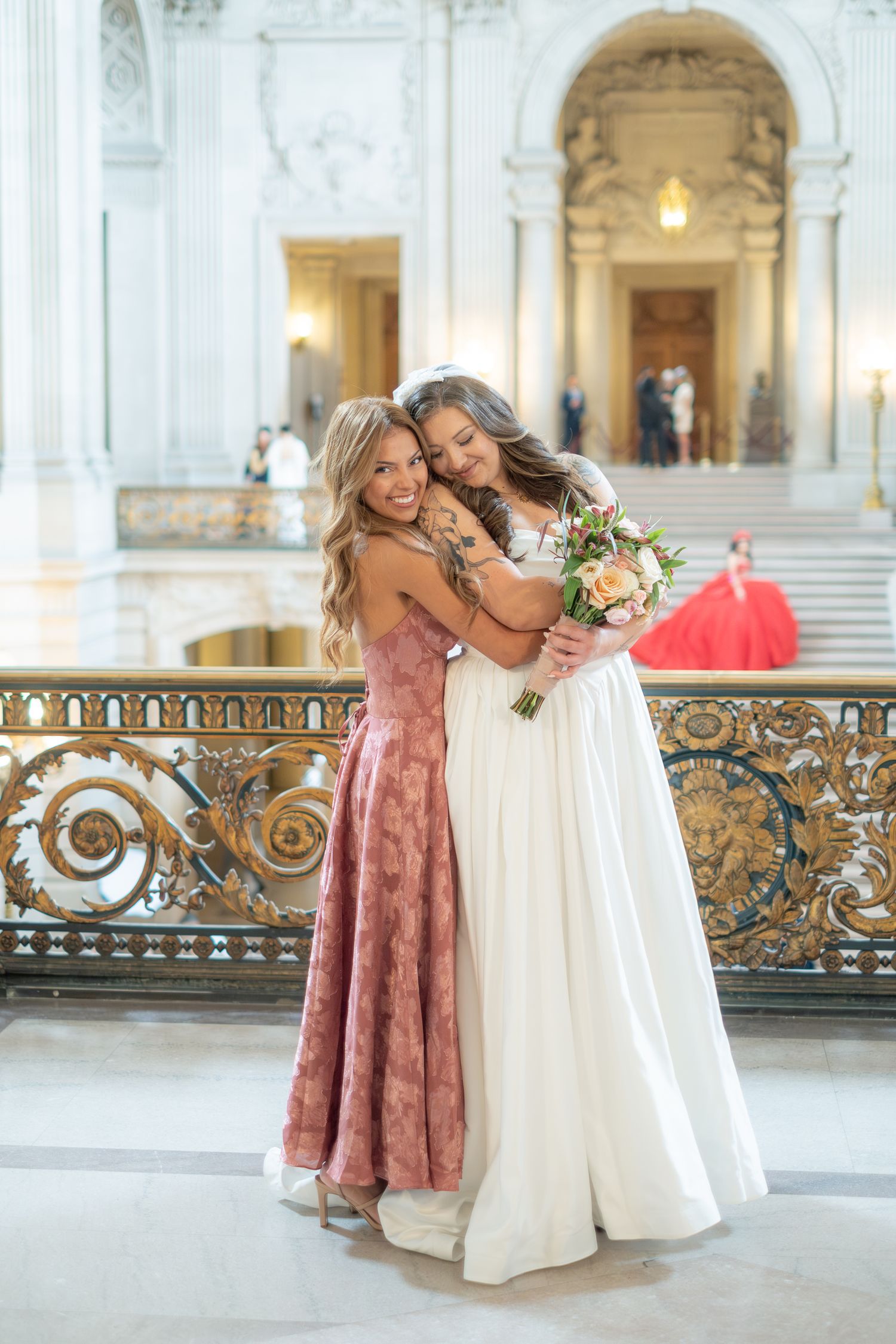 Bride in white gown hugs bridesmaid in pink dress, holding bouquet at elegant San Francisco City Hall.