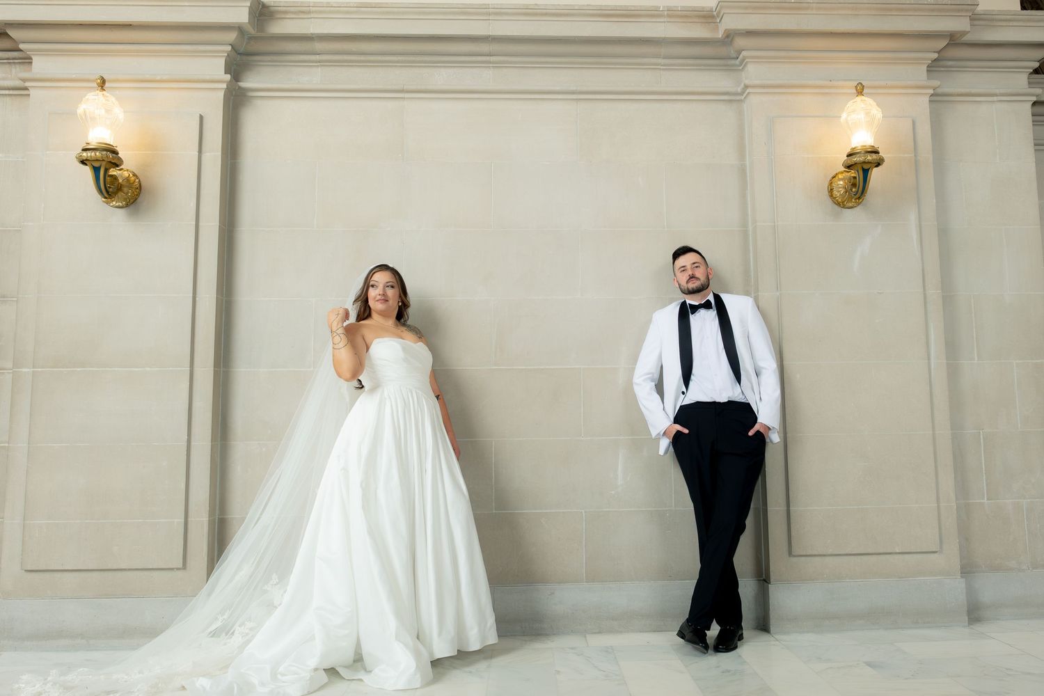 Bride in white ballgown and groom in tuxedo pose apart against elegant marble wall with gold sconces.