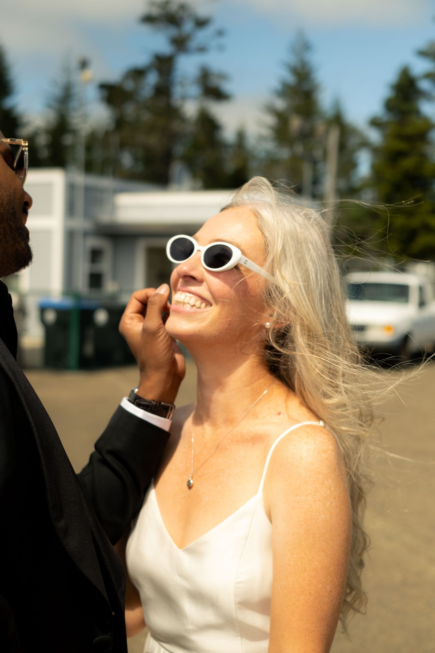 Smiling blonde bride in white dress and sunglasses laughing outdoors on sunny wedding day.
