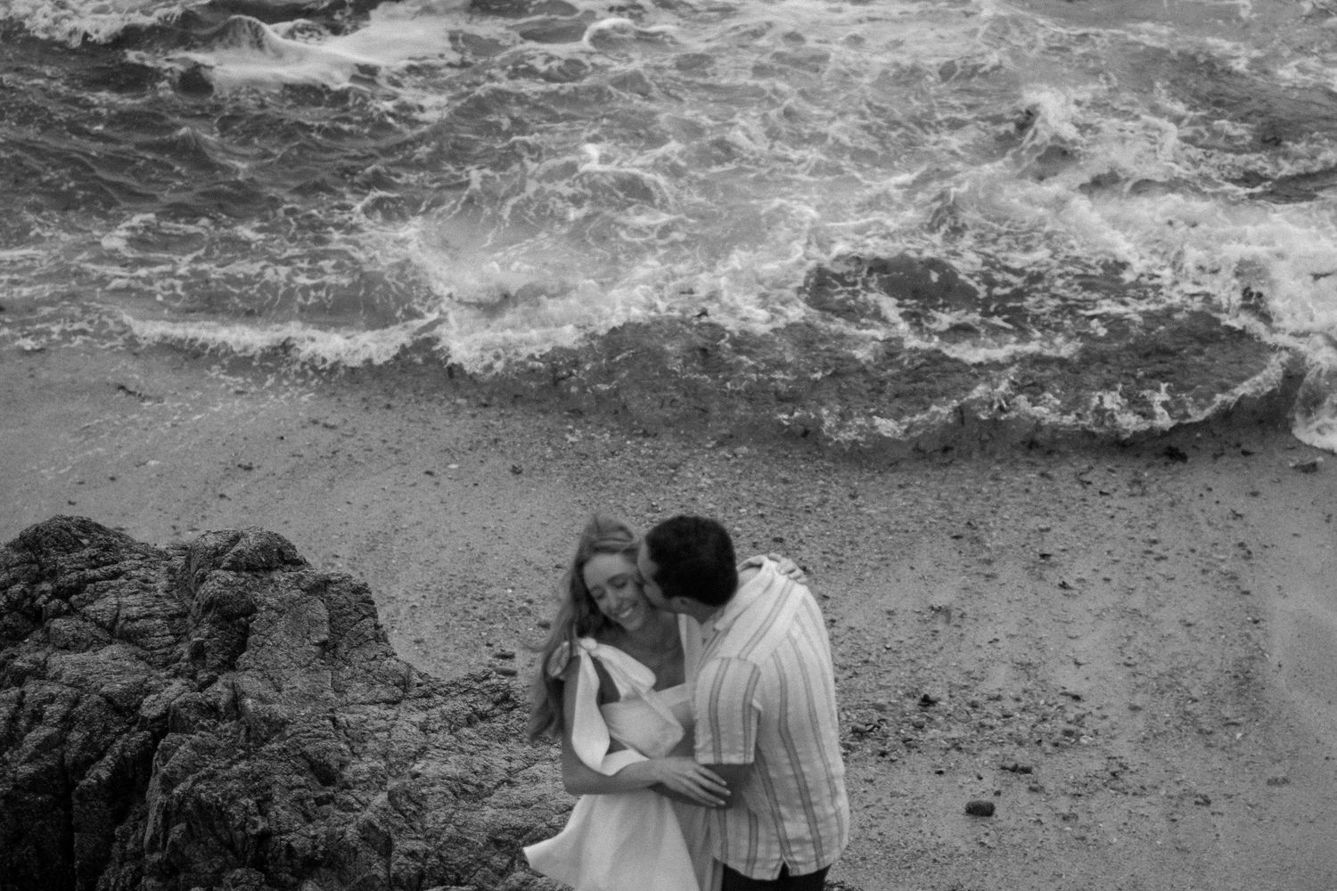 Black and white photo of a couple sharing a tender moment on a rocky beach with ocean waves.