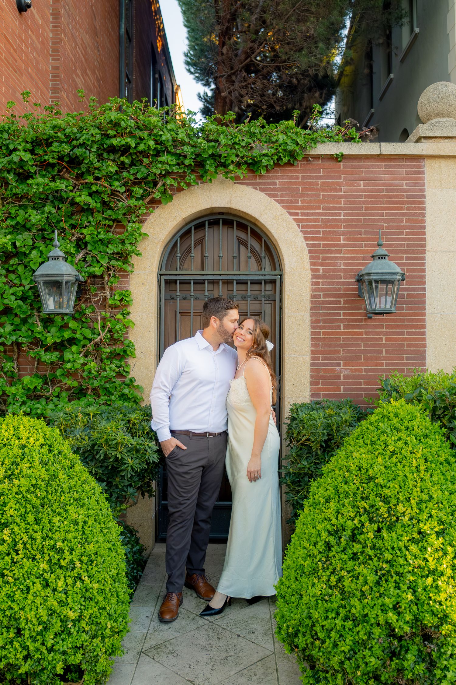 A couple shares a romantic kiss in front of an arched brick doorway surrounded by lush greenery and manicured hedges.