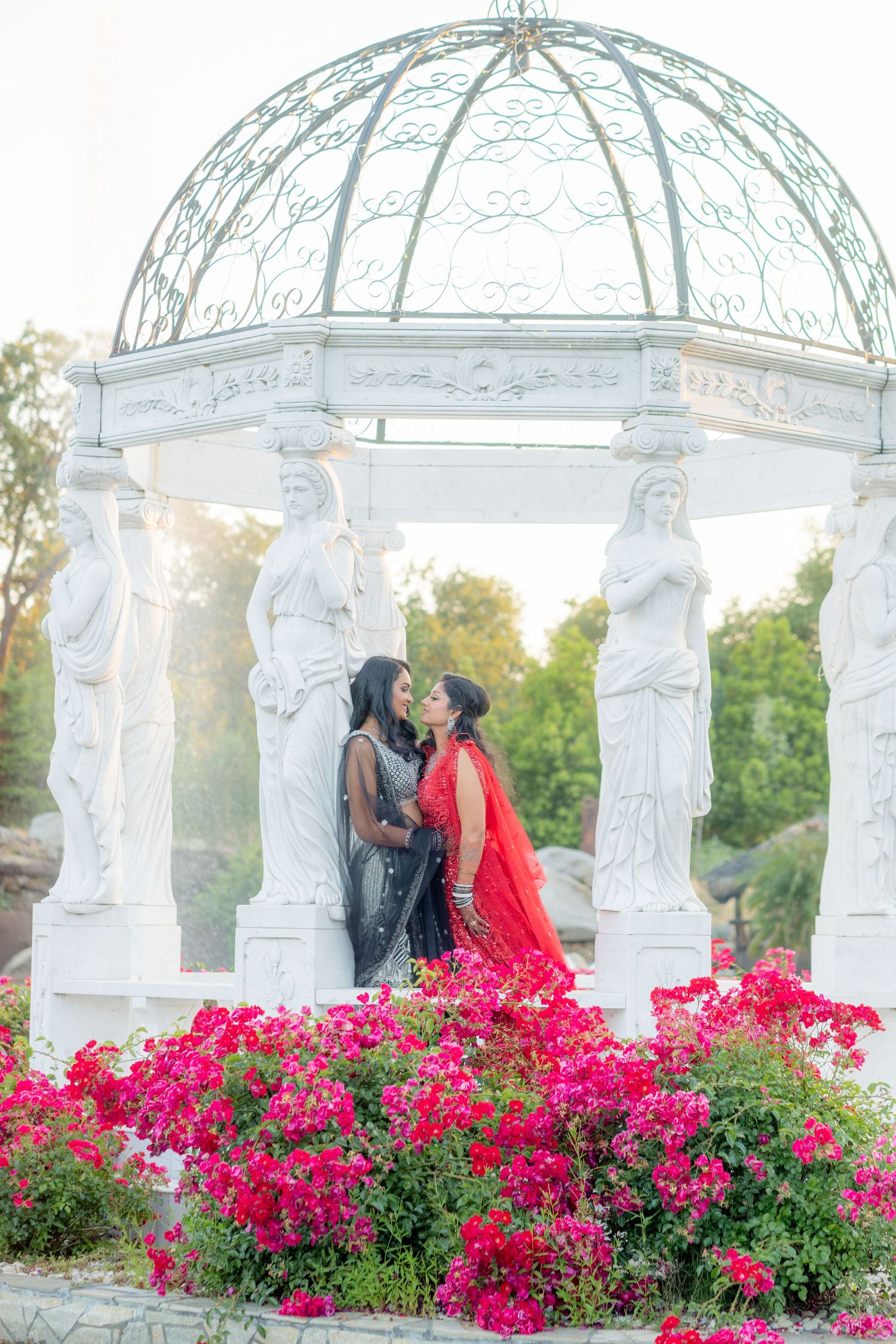 Two women in red and grey Indian lehengas share an intimate moment under a white ornate gazebo with pink flowers.
