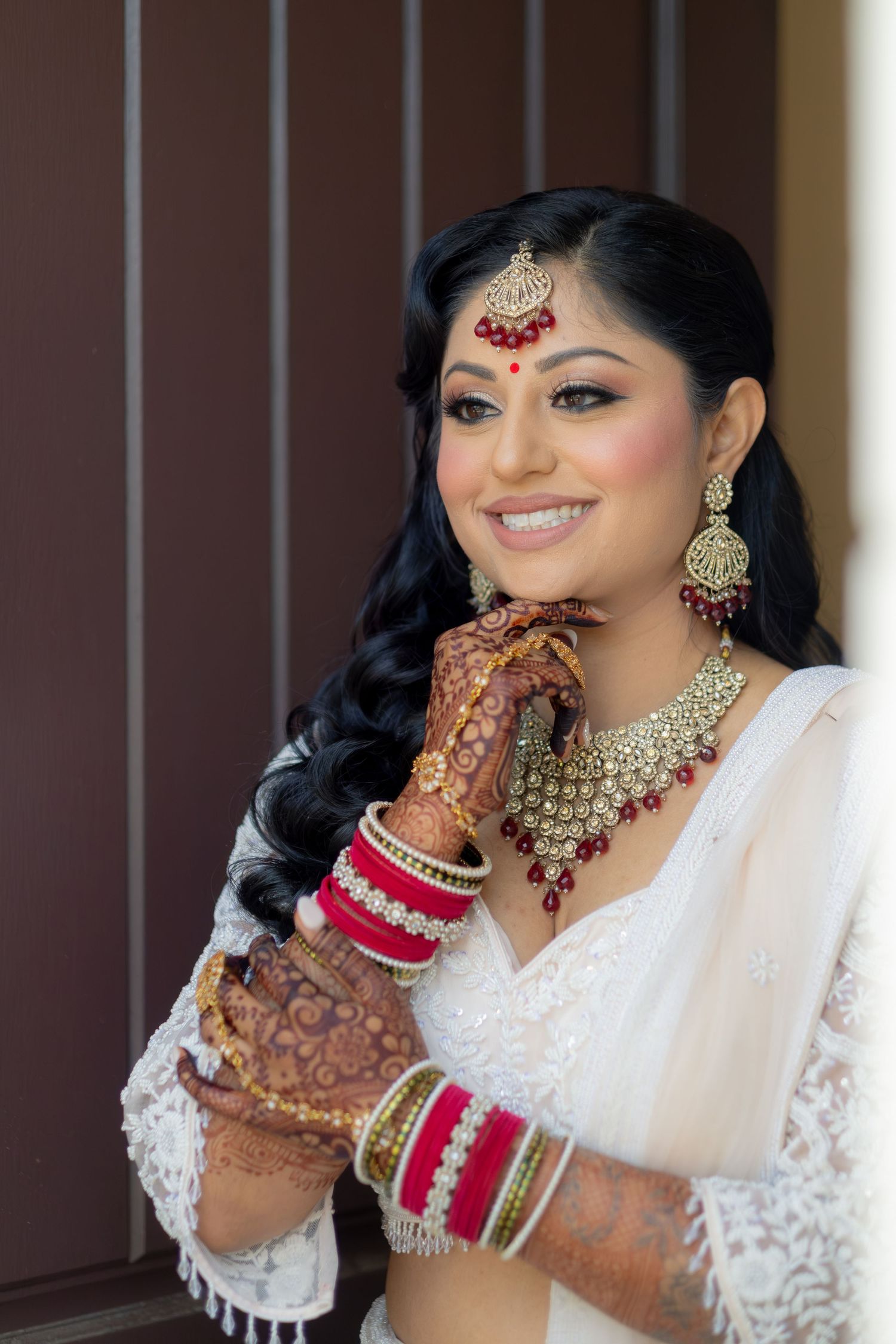Indian bride smiling in white saree with red jewelry, bangles, and mehndi on wedding day.