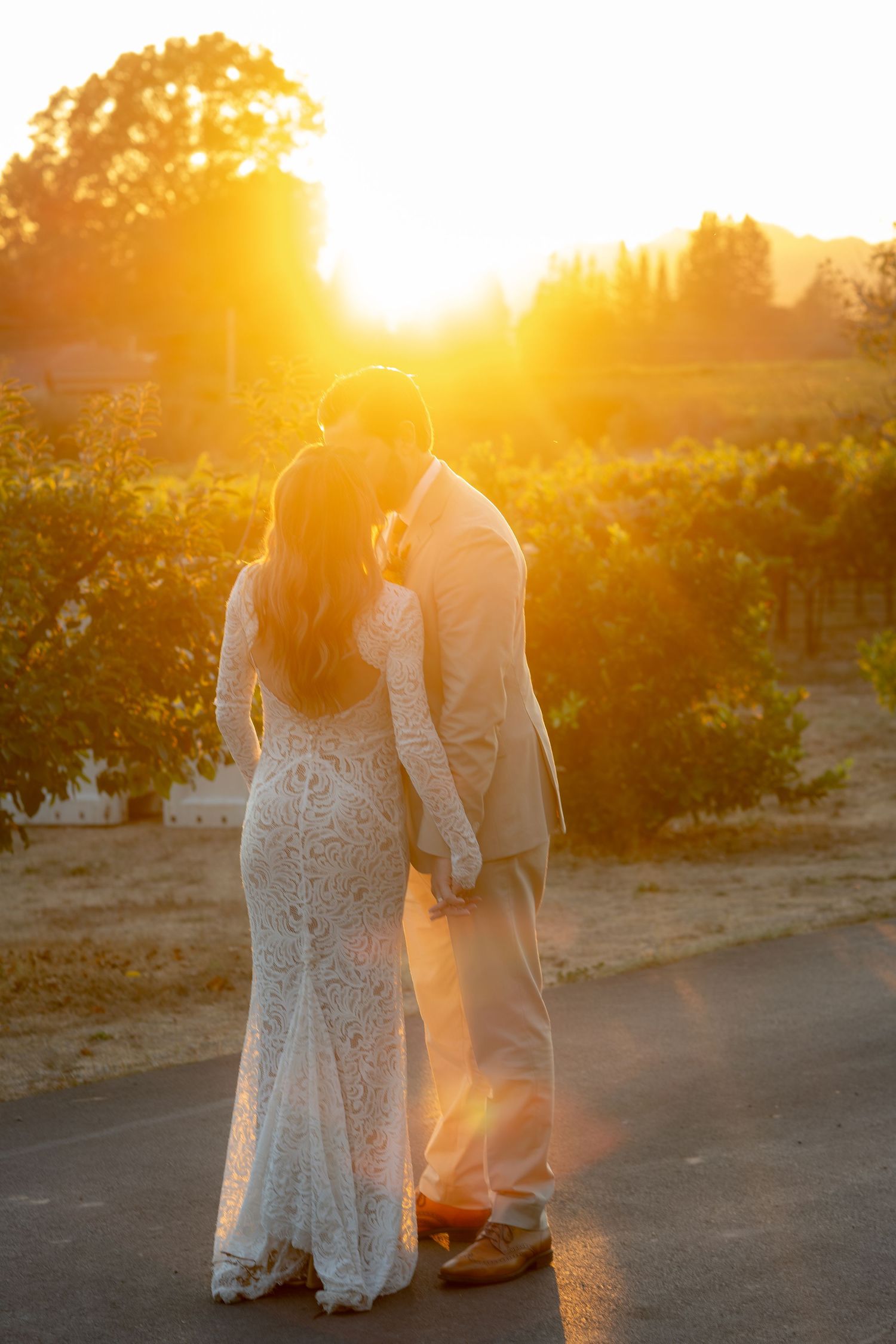 Bride and groom silhouetted against a golden sunset overlooking a lush vineyard on their wedding day.