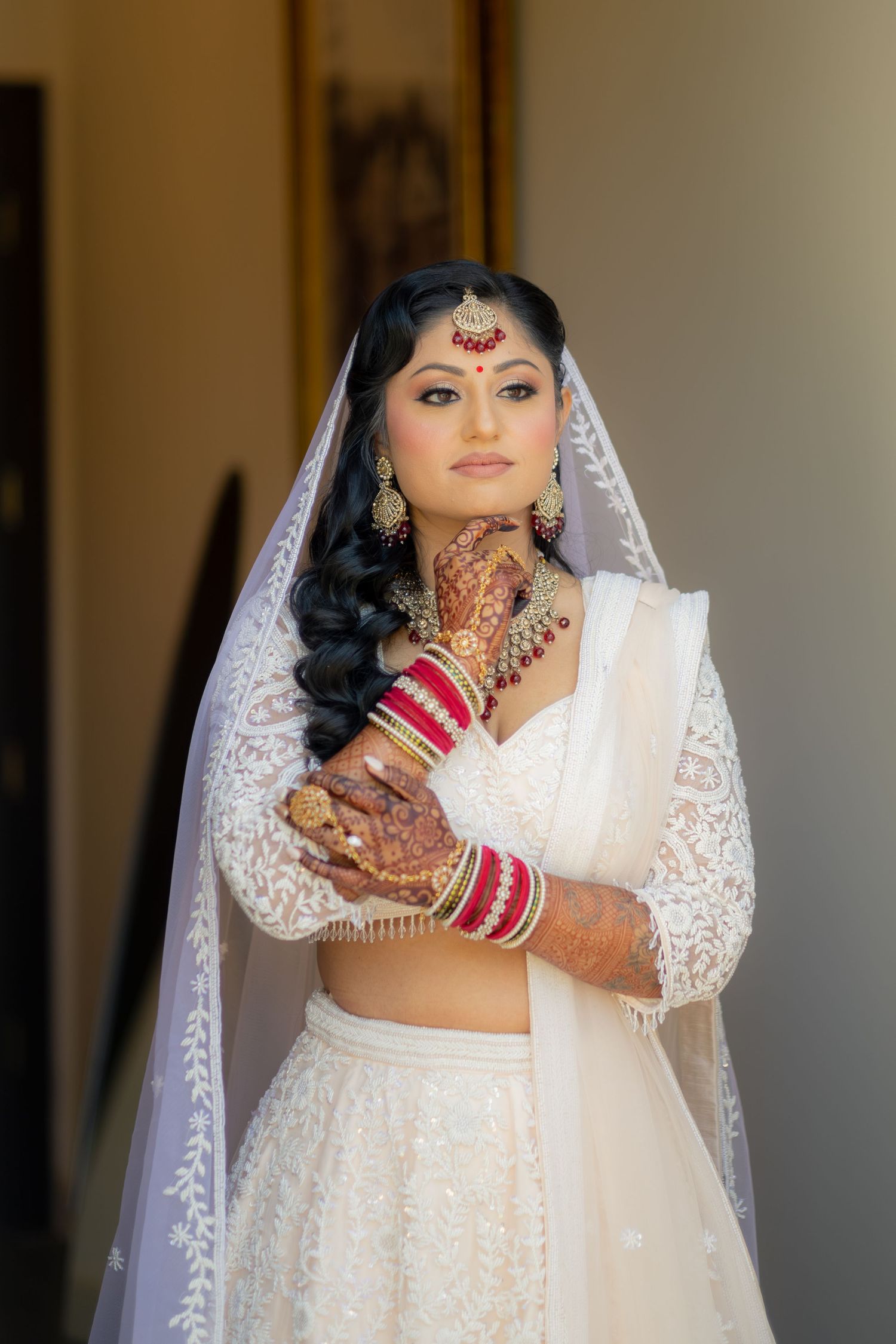 Indian bride in white lace lehenga with red bangles, mehndi, and bridal jewelry poses elegantly with veil.