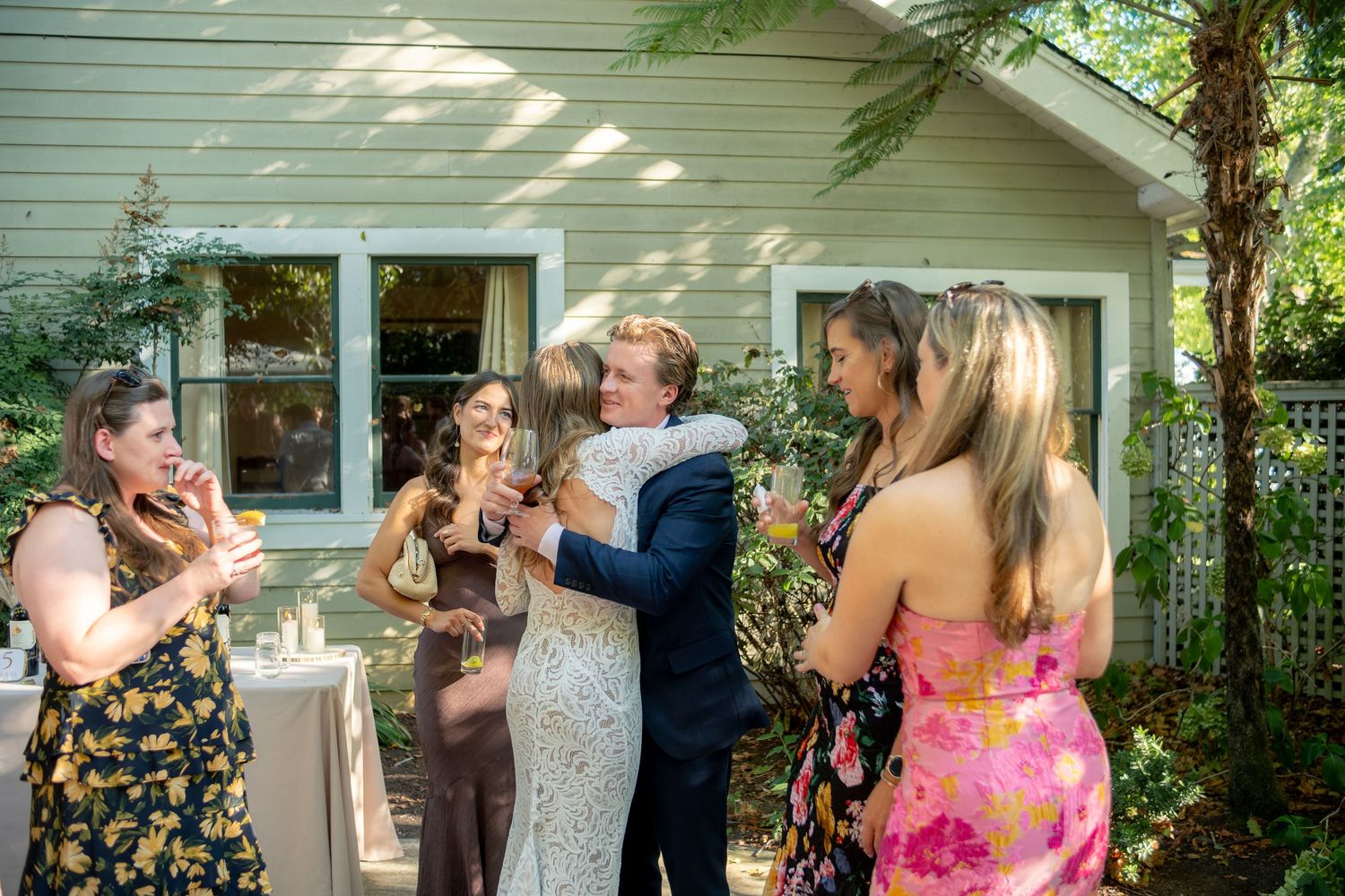 Bride in white gown hugs guest at outdoor wedding reception while smiling friends gather around in colorful dresses.