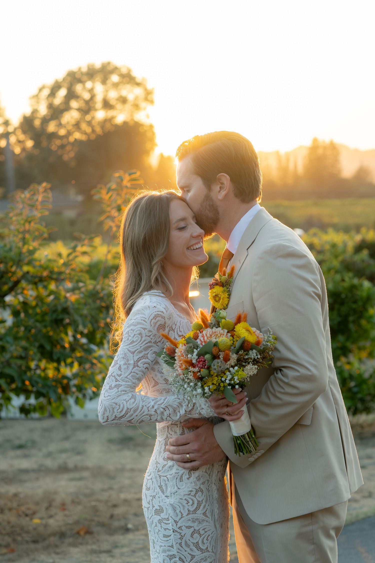 Groom kisses bride holding colorful bouquet during golden hour at an outdoor vineyard wedding.