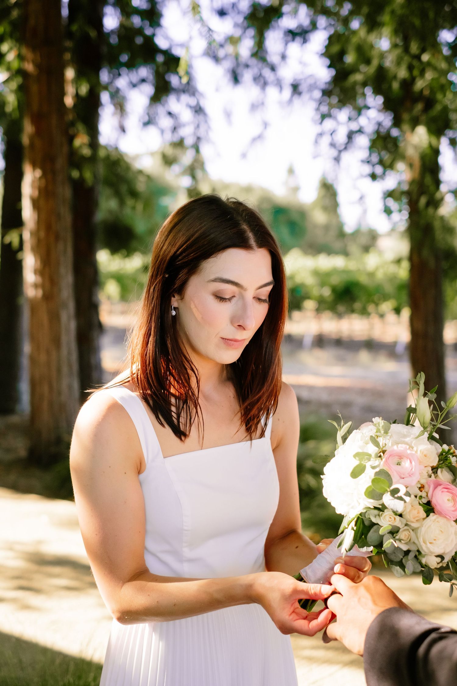 Bride in white dress receiving wedding ring from groom during outdoor ceremony in vineyard setting.