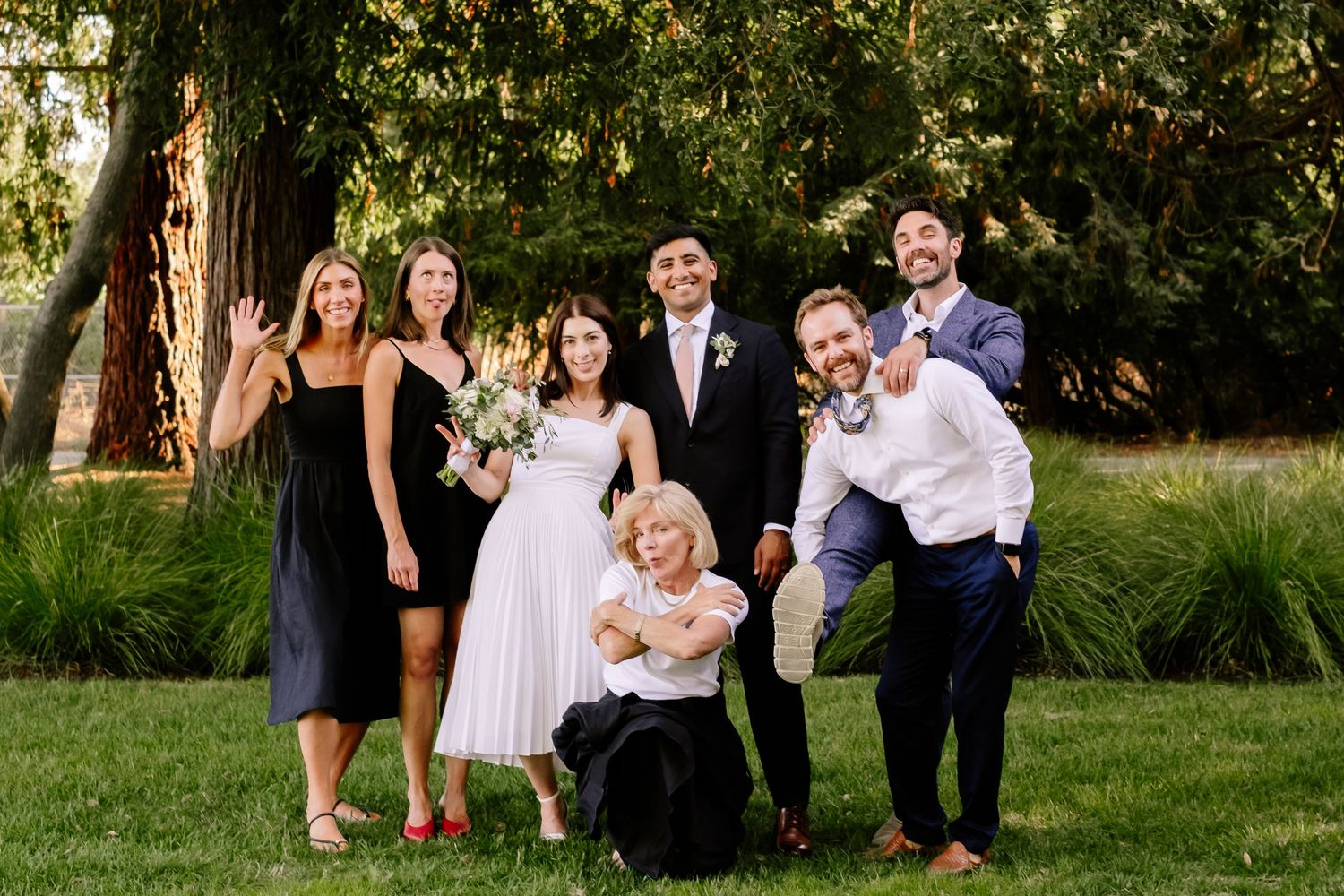 Wedding party posing joyfully outdoors on green lawn with trees, bride in white dress holding bouquet with guests.