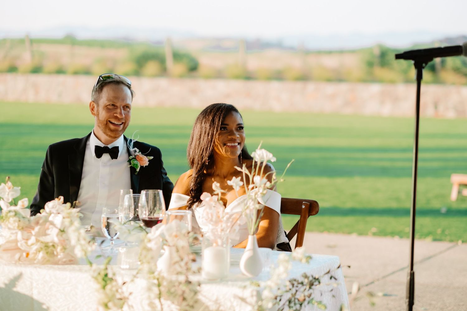 A laughing bride and groom in formal attire sit at their sweetheart table during outdoor wedding reception speeches.