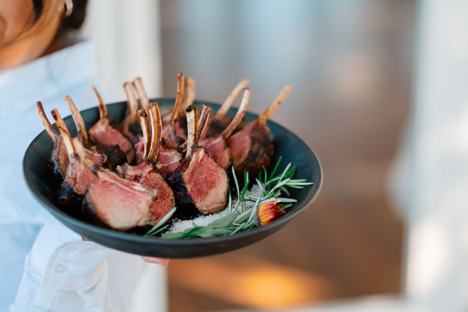 A server holds a dark plate of elegant frenched lamb chops garnished with fresh rosemary at a wedding reception.