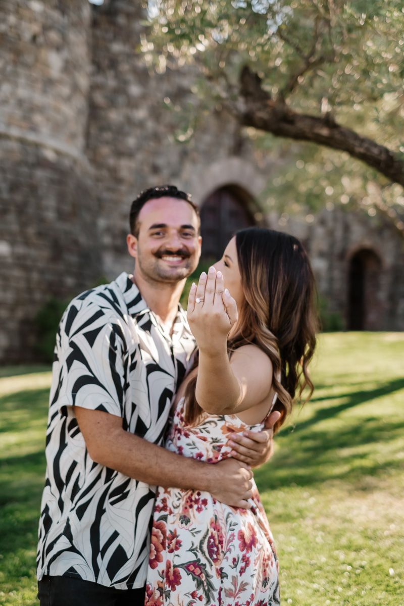 Smiling man embracing woman showing off her engagement ring in front of a stone castle.