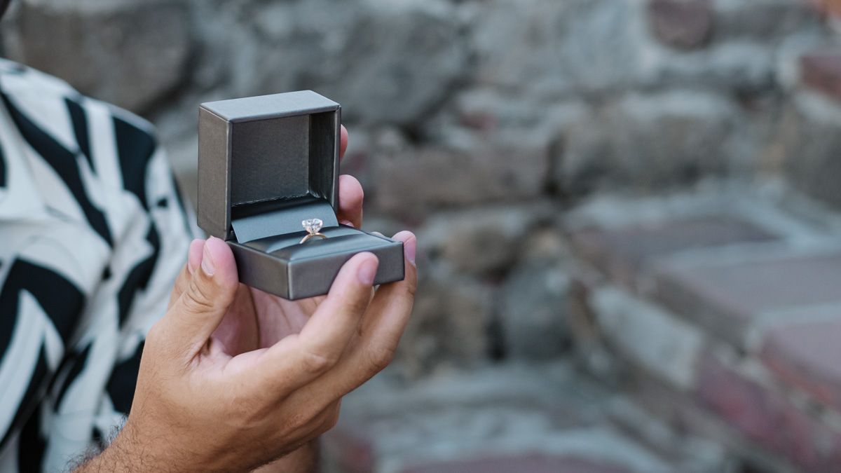 Hands holding an open gray jewelry box with a diamond engagement ring against a stone wall background.