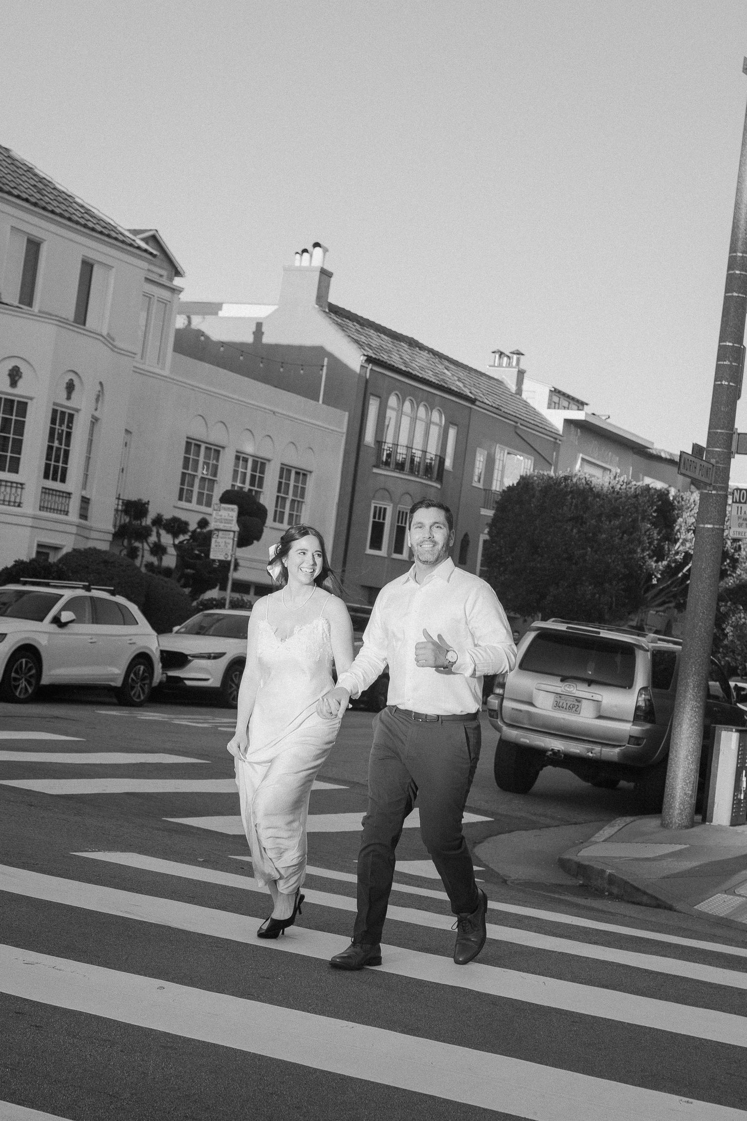 Black and white photo of a couple holding hands while crossing a street crosswalk in an urban neighborhood.