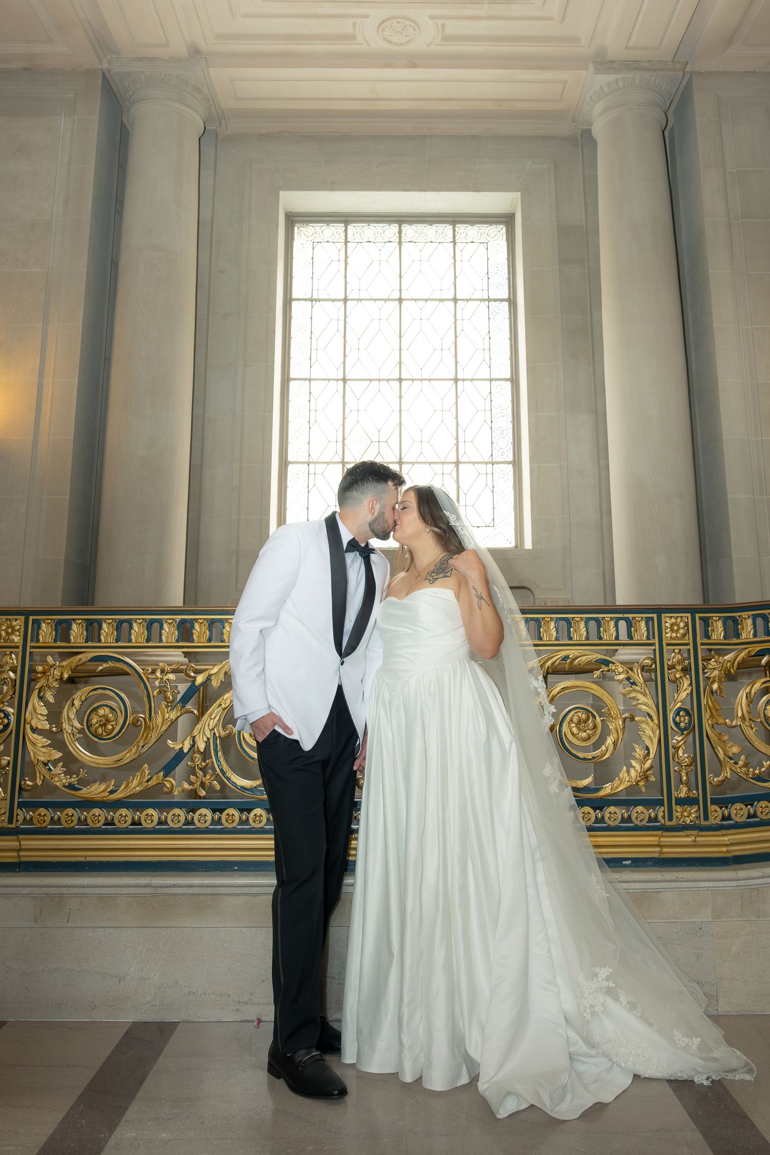 Newlyweds share a kiss in front of a sunlit stained glass window at San Francisco City Hall.