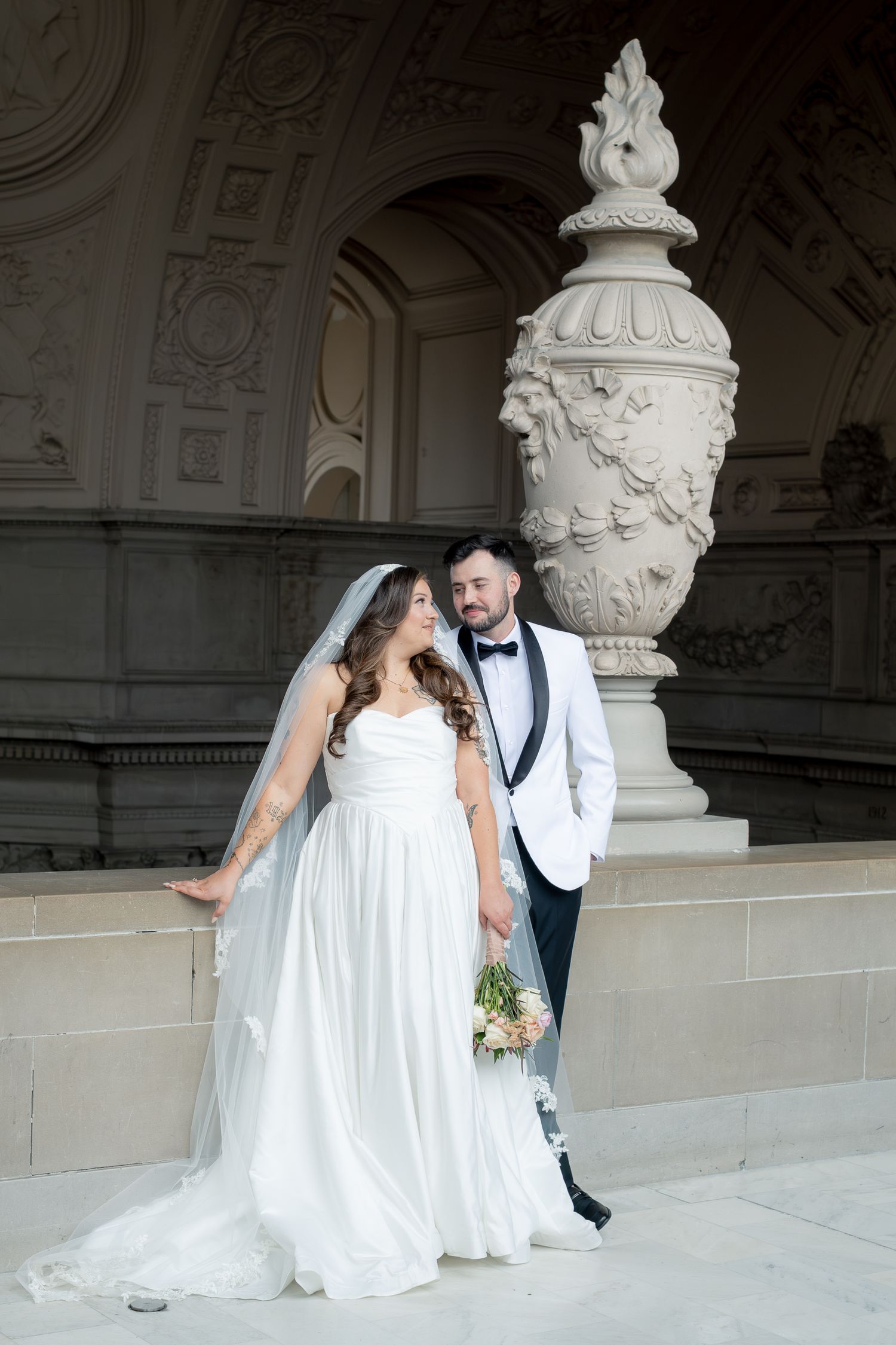 Bride and groom posing at ornate stone building, she in white gown with veil, he in white tuxedo jacket.
