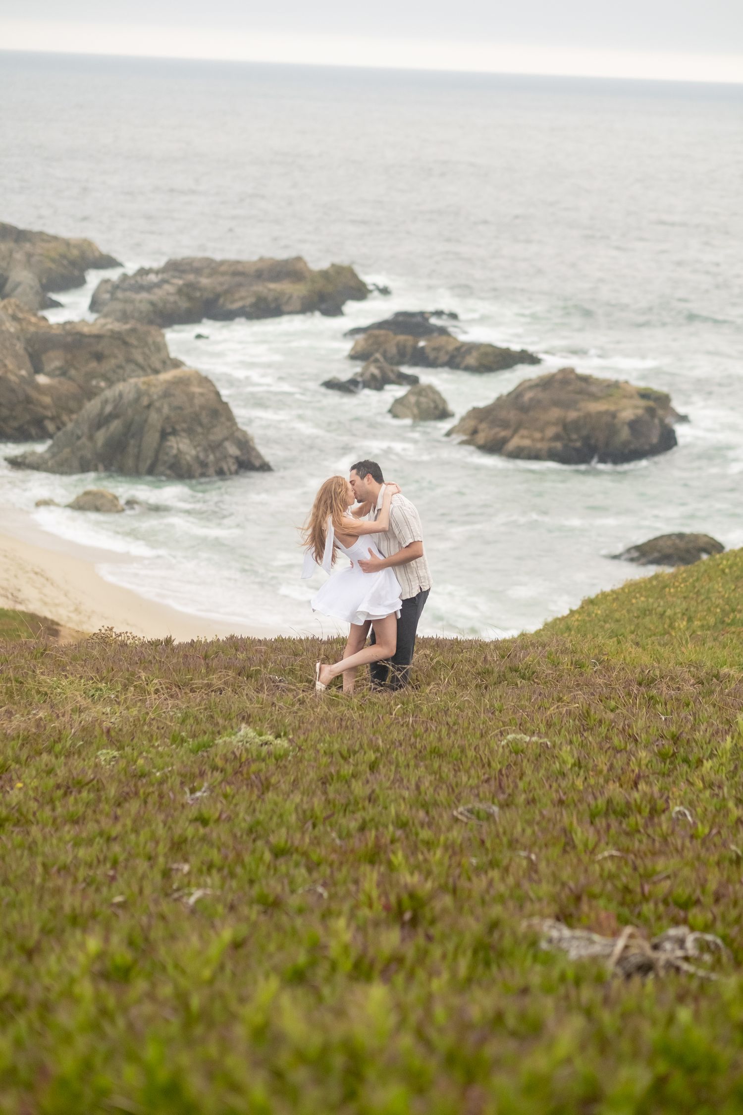Couple kissing on a green coastal hillside with rocky beach and ocean waves below.