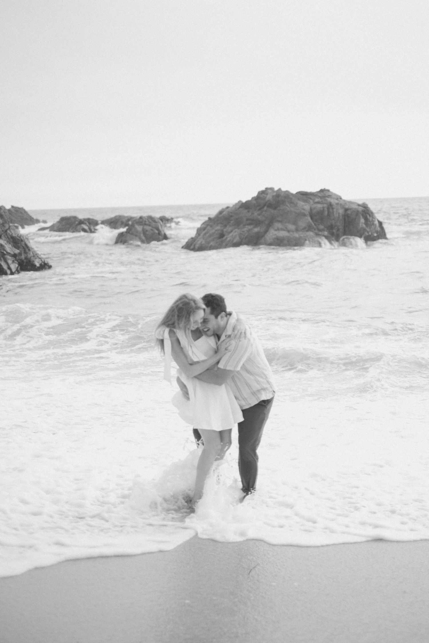 Black and white photo of a couple kissing in ocean waves with rocky outcrops behind them.