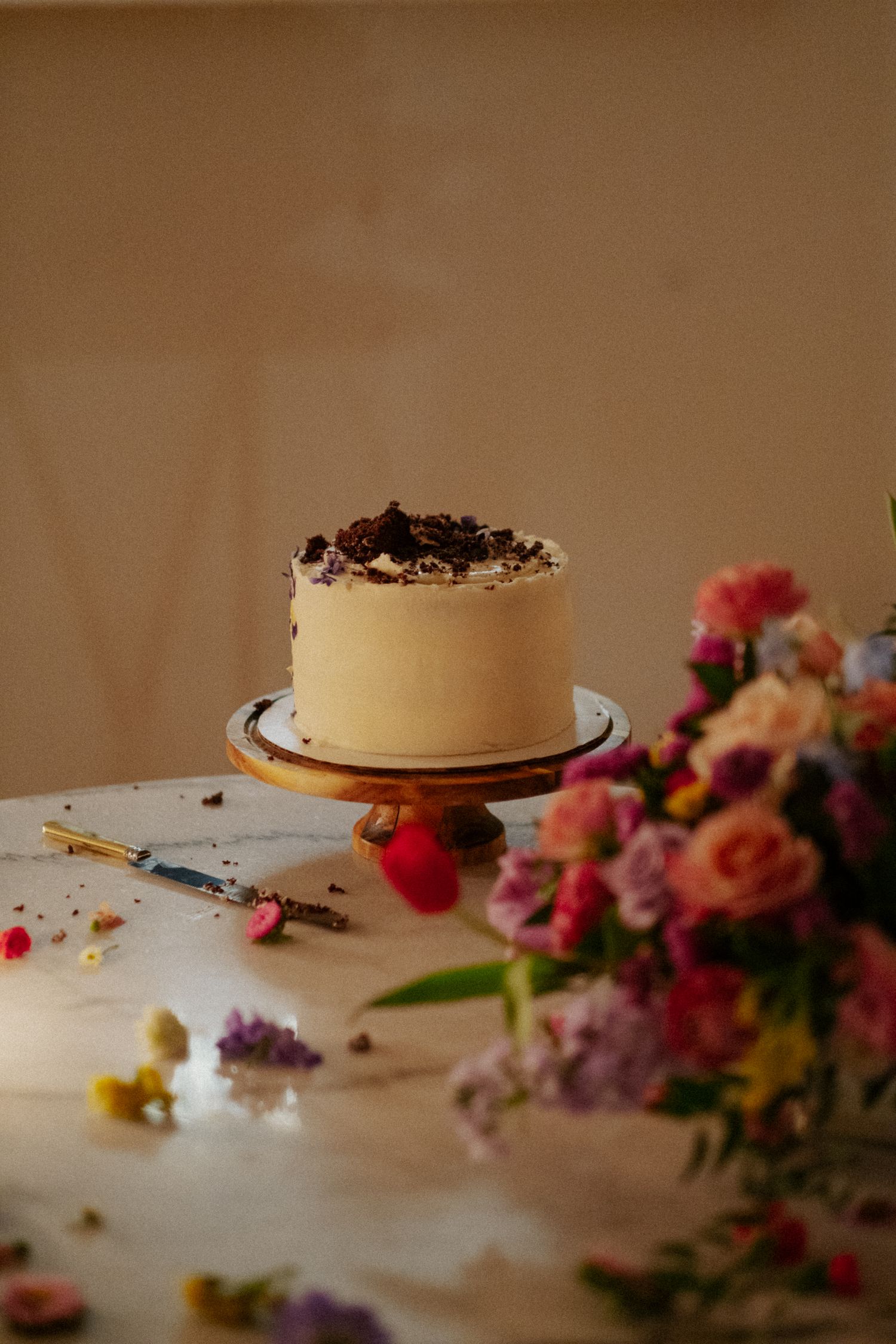 Elegant single-tier white wedding cake with chocolate topping on wooden stand beside colorful florals.