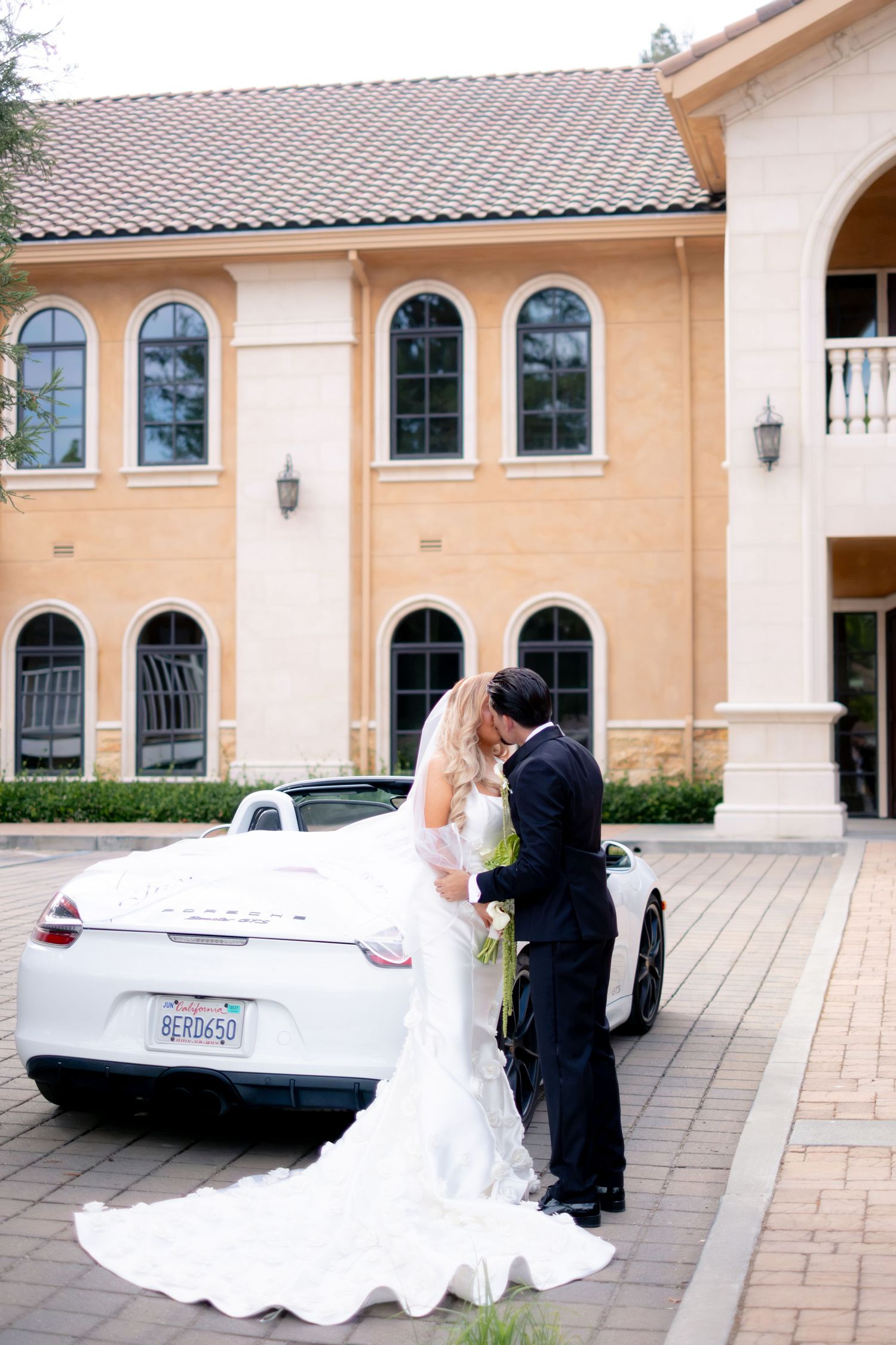 Bride and groom kissing beside white Porsche convertible outside elegant venue.