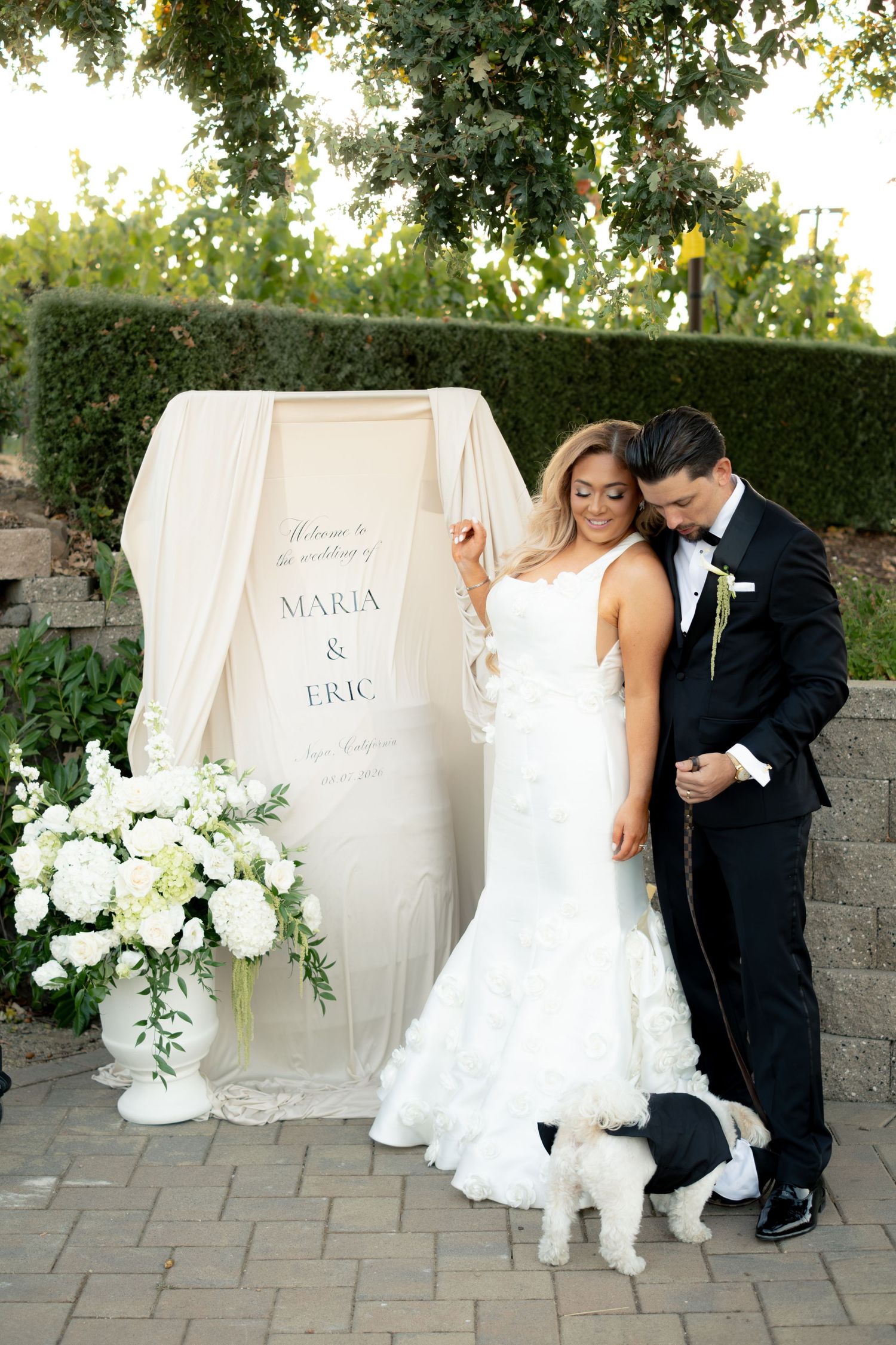 Bride and groom pose with their white dog beside a welcome sign reading Maria and Eric.