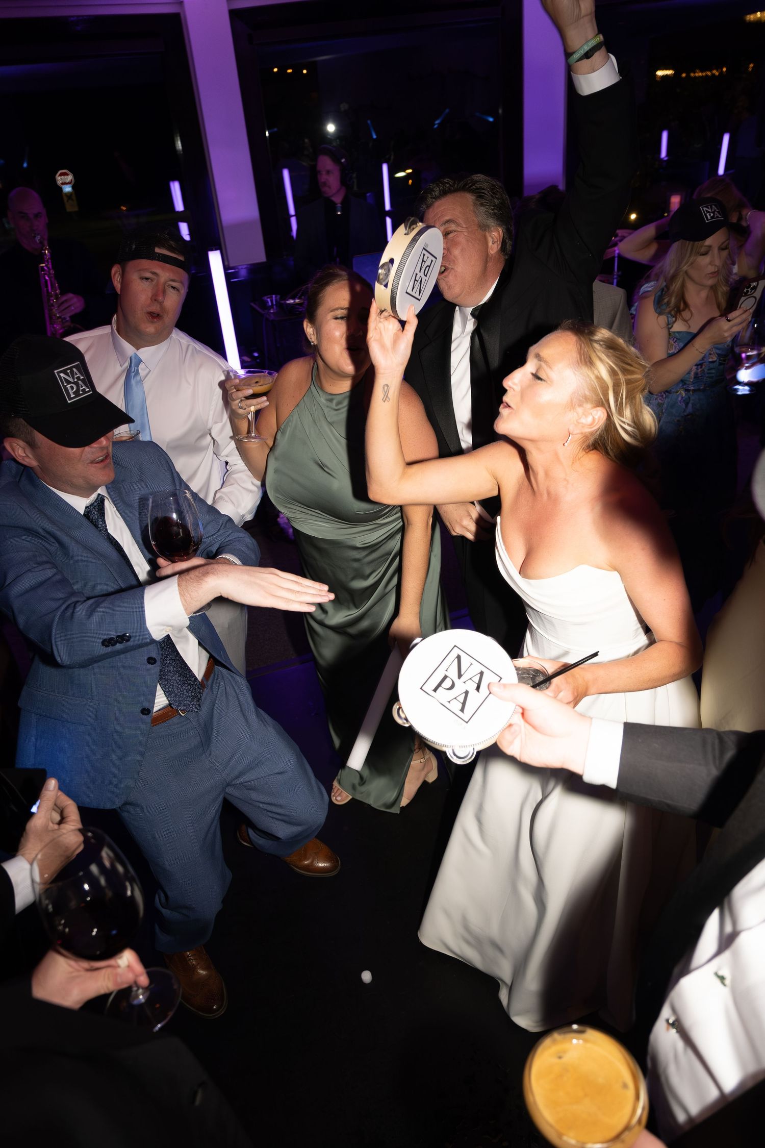 Bride in white gown dancing with guests holding tambourines at energetic indoor wedding reception.