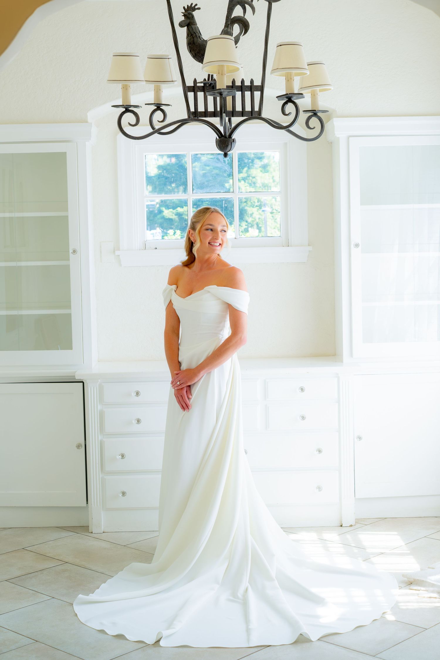 Radiant bride poses in elegant off-shoulder white wedding gown beneath ornate chandelier in bright room.