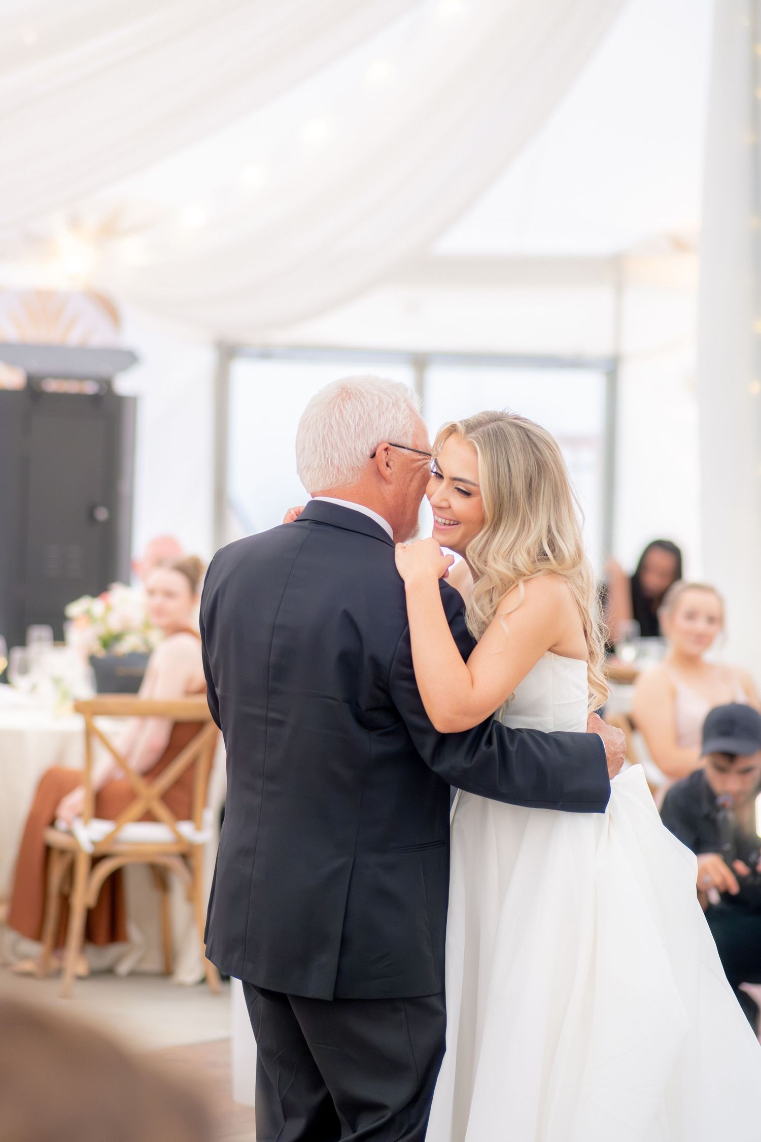 Bride sharing tender father-daughter dance inside elegant white draped wedding reception venue.