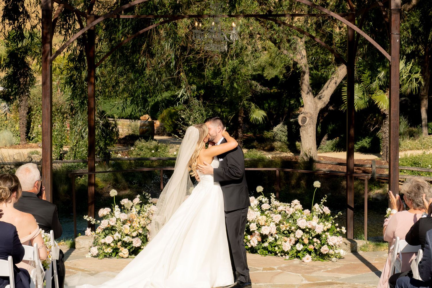 Bride and groom share first kiss under floral arch altar beside a pond during outdoor wedding ceremony.