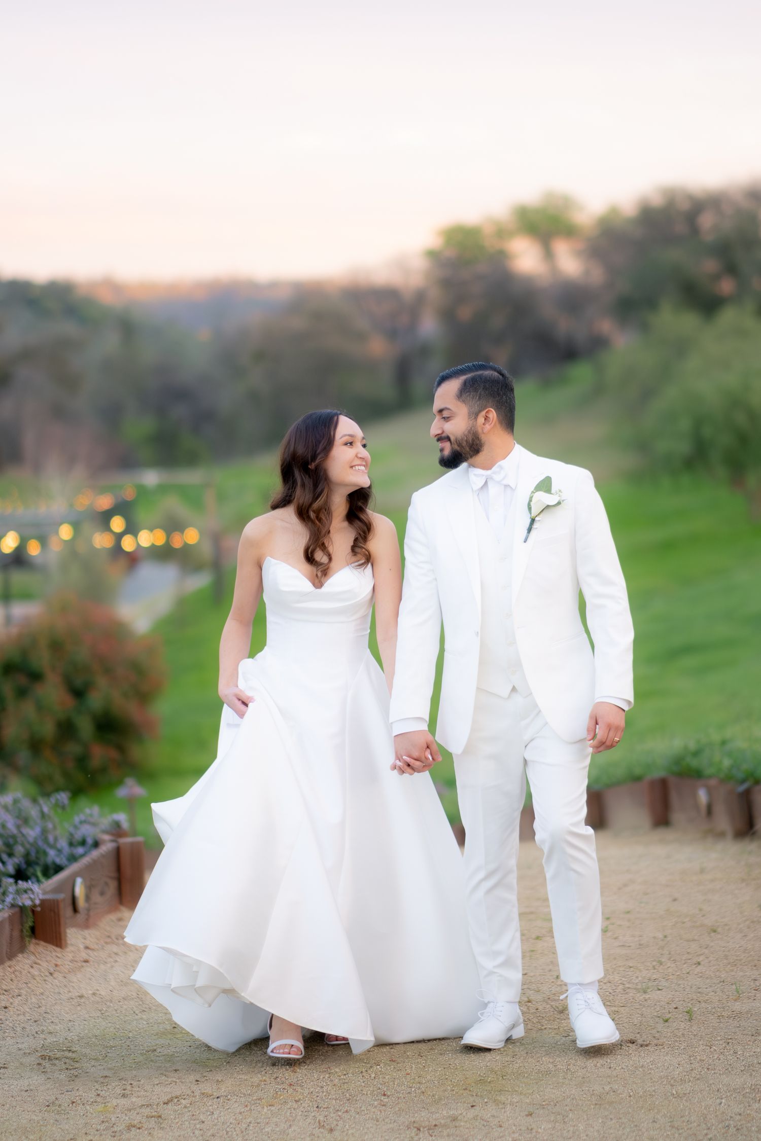 Bride and groom in white attire hold hands walking at sunset with green hills and string lights in background.