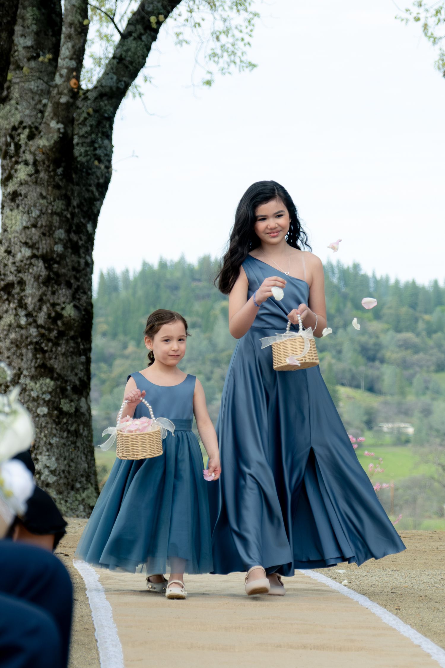 Two flower girls in blue dresses carry wicker baskets down an outdoor wedding aisle.