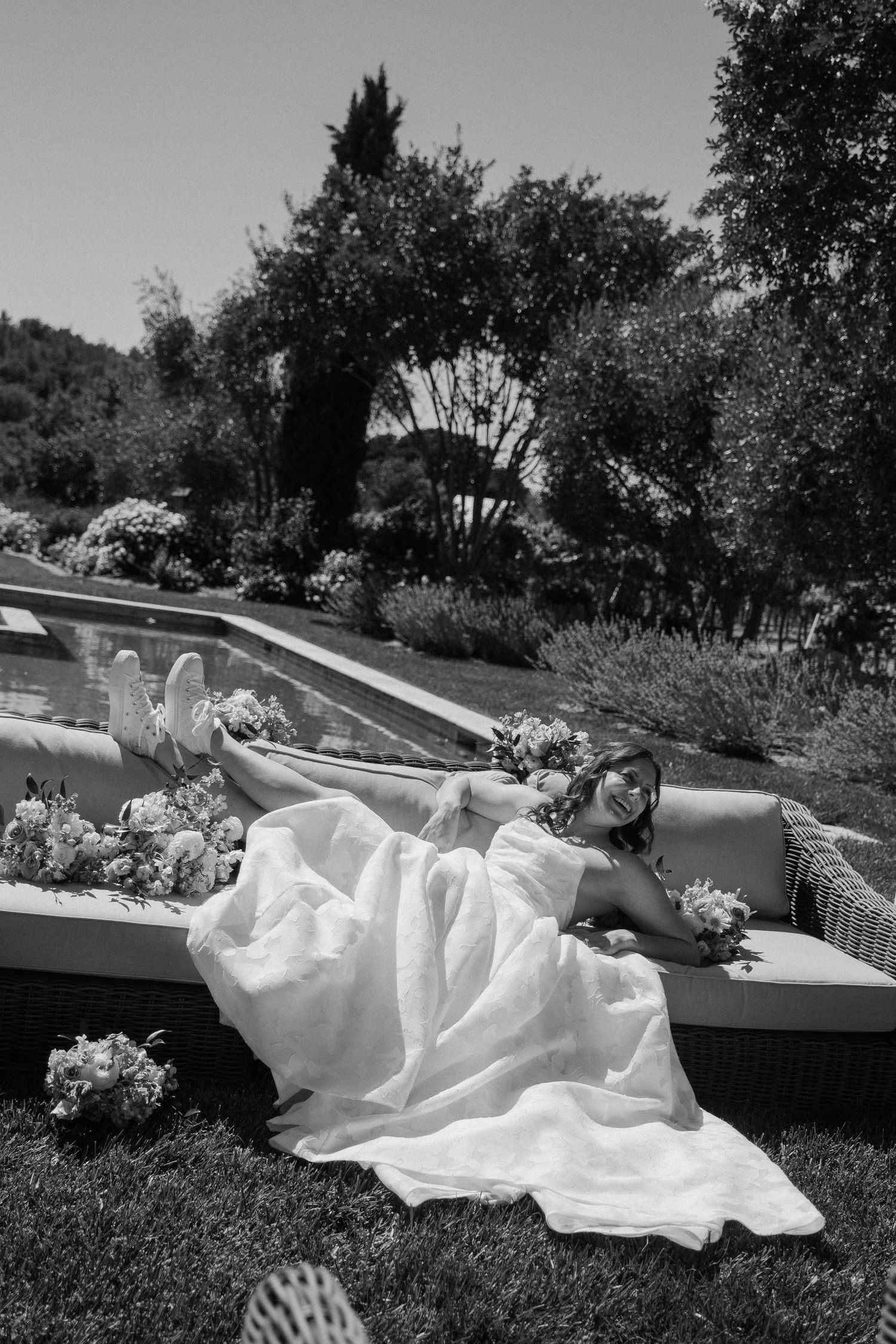 Black and white photo of a bride in a white gown relaxing on outdoor furniture, holding bouquets, surrounded by lush garden greenery.