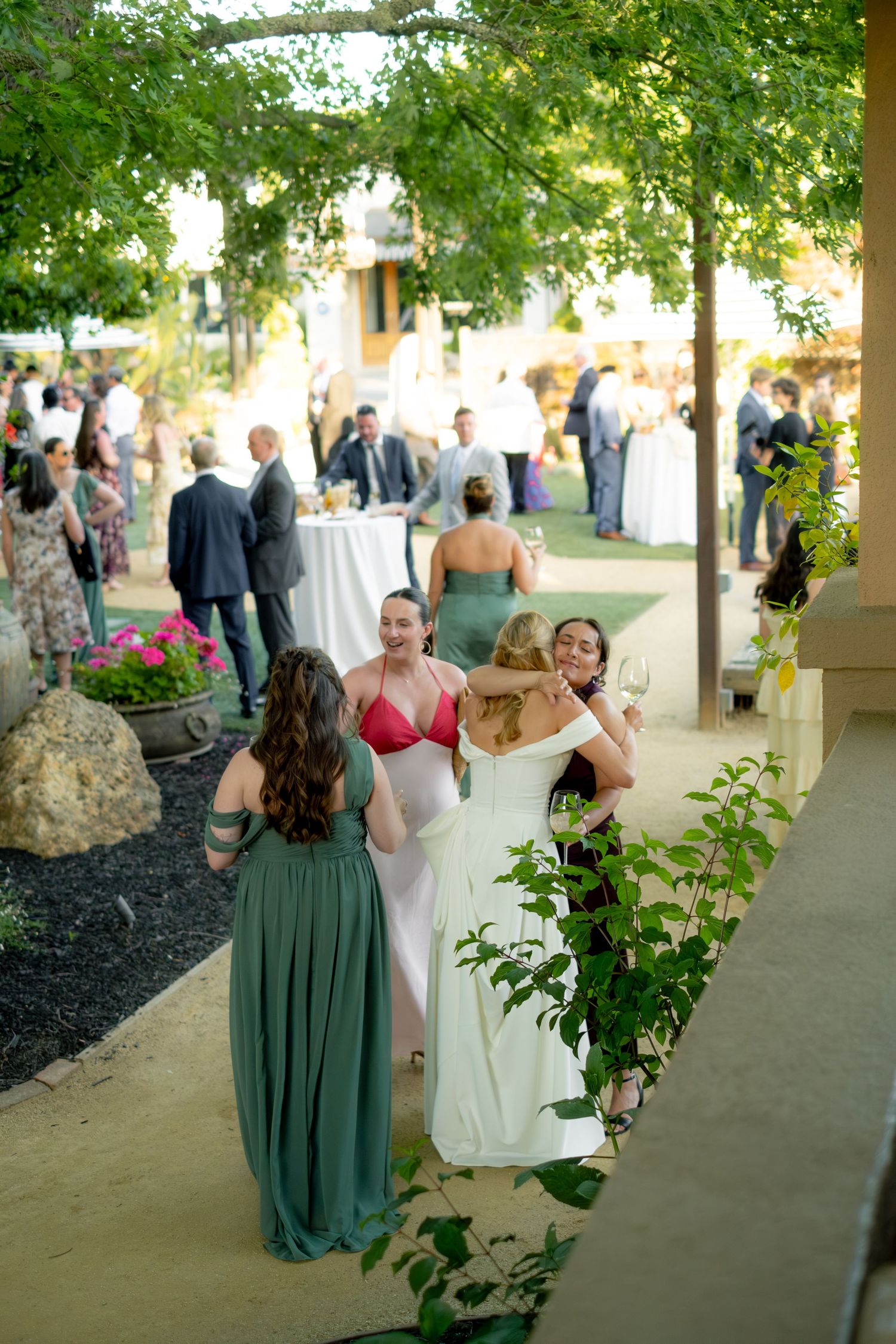 Wedding guests mingling outdoors under green vine-covered pergola with women in elegant dresses enjoying cocktail hour.