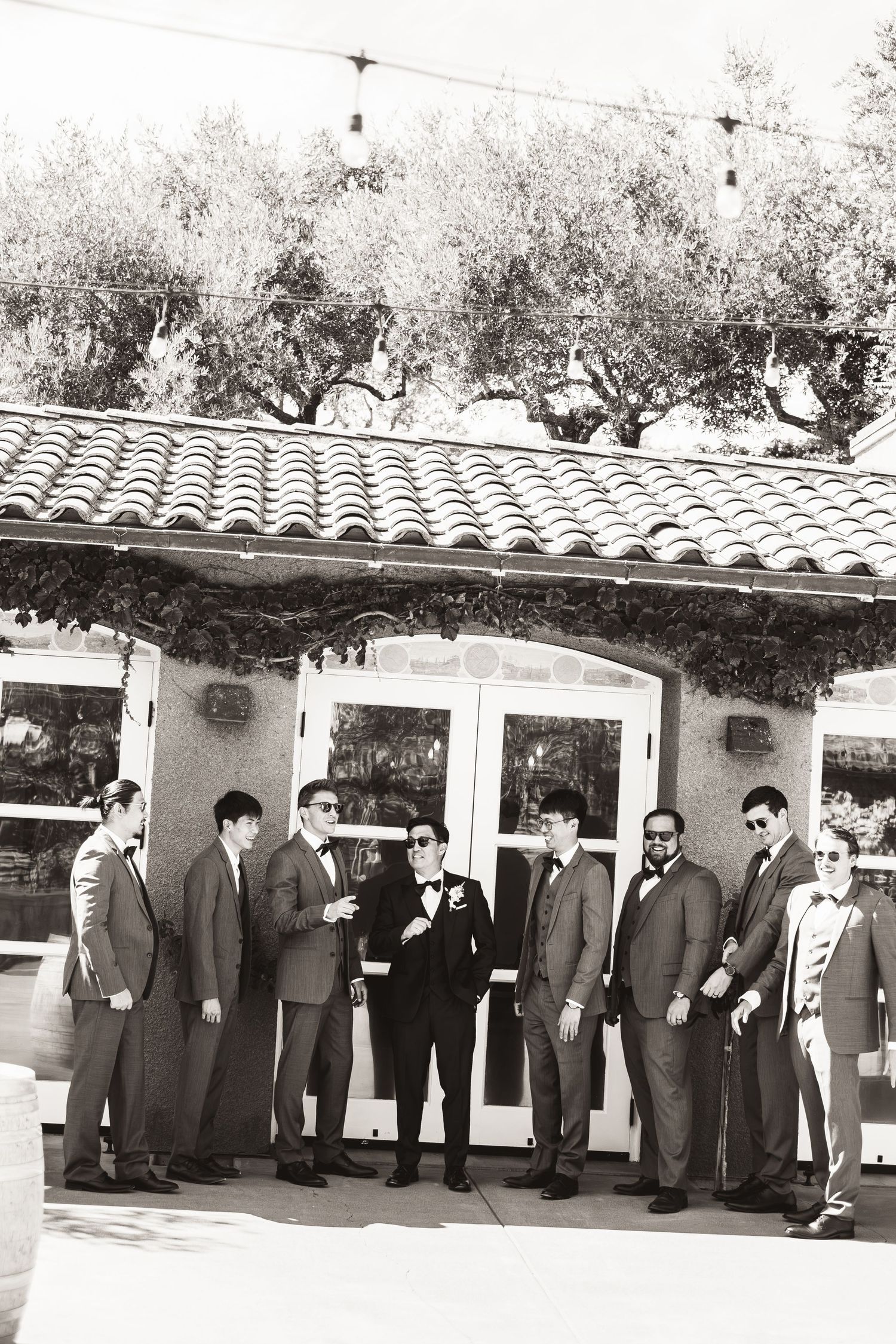 Groom and groomsmen in gray suits pose in black and white photo outside Spanish-style venue.