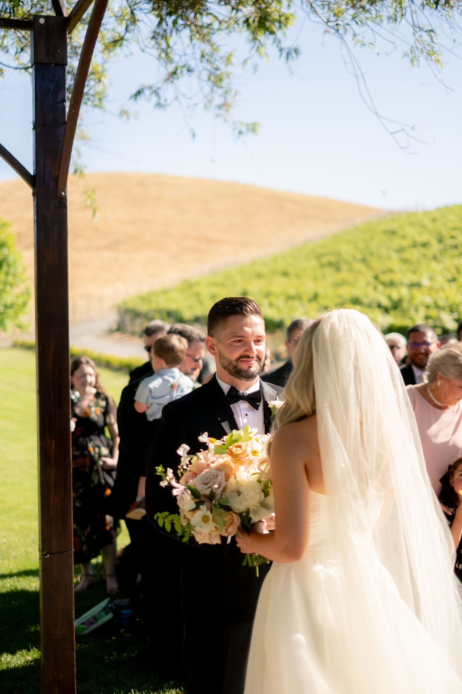 Groom smiling at bride during outdoor vineyard wedding ceremony with rolling hills in background.