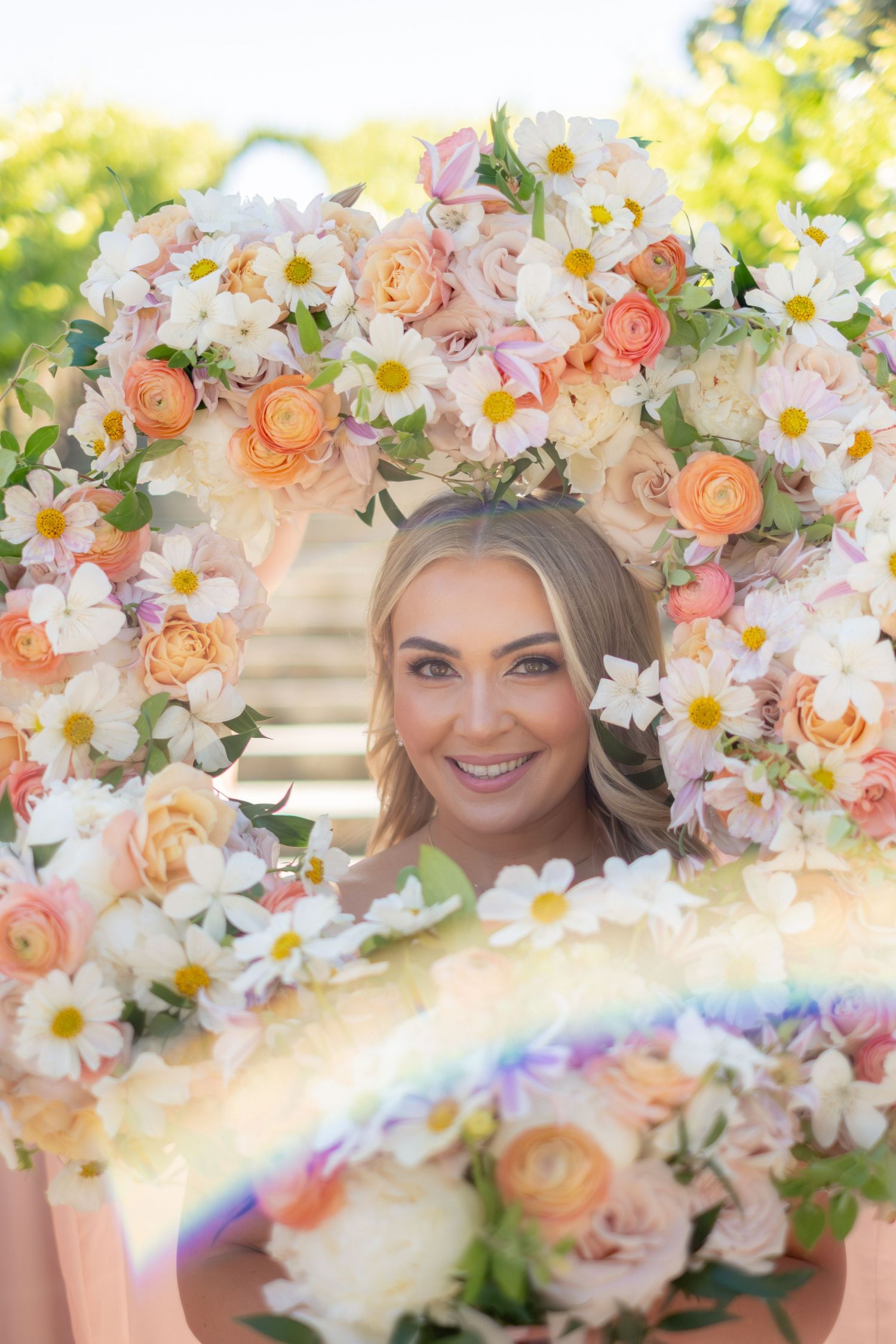 Smiling bride peeks through a large circular wreath of peach roses, daisies, and ranunculus blooms.