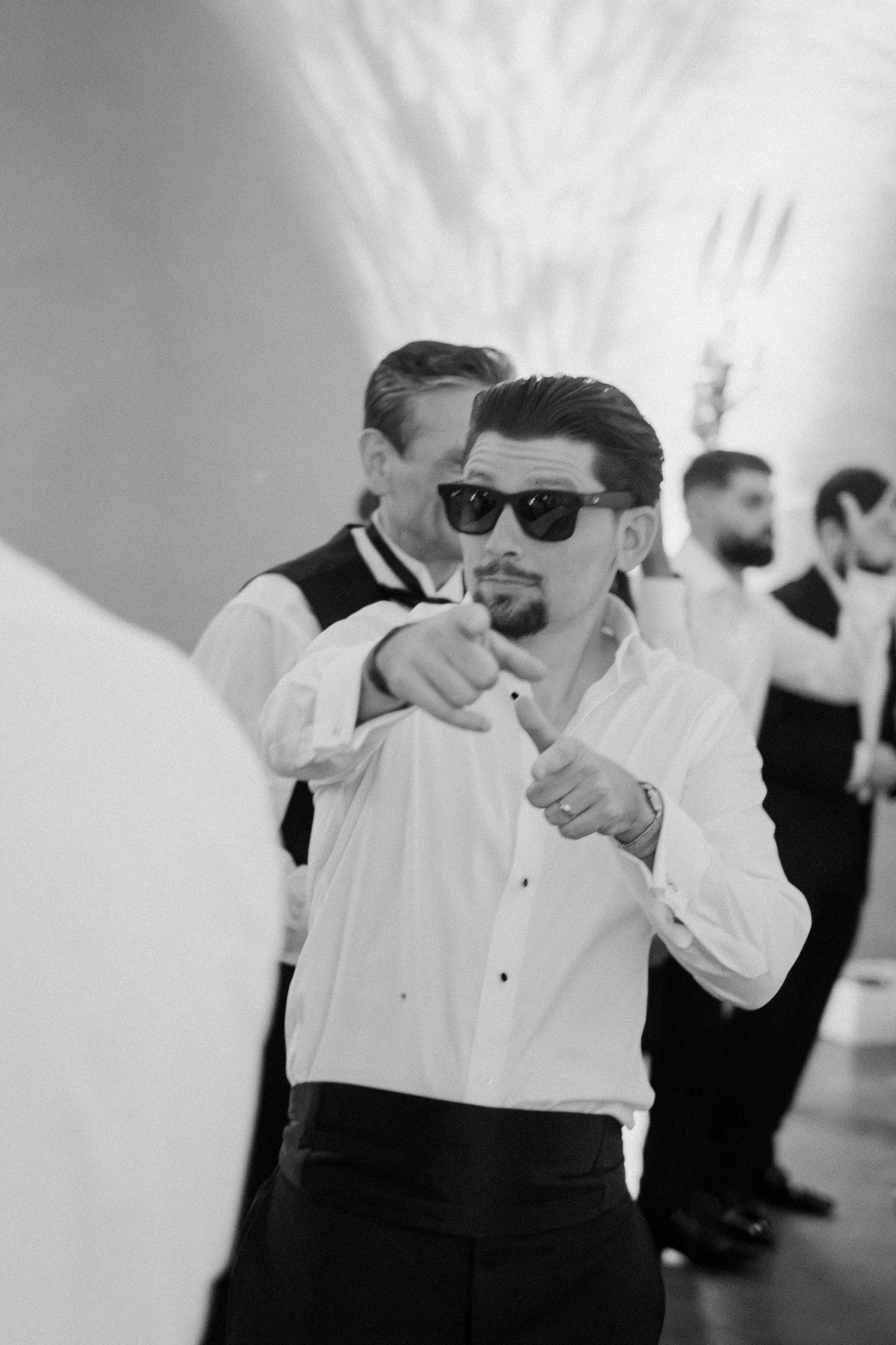 Black and white photo of groomsman in sunglasses pointing at camera during wedding reception.