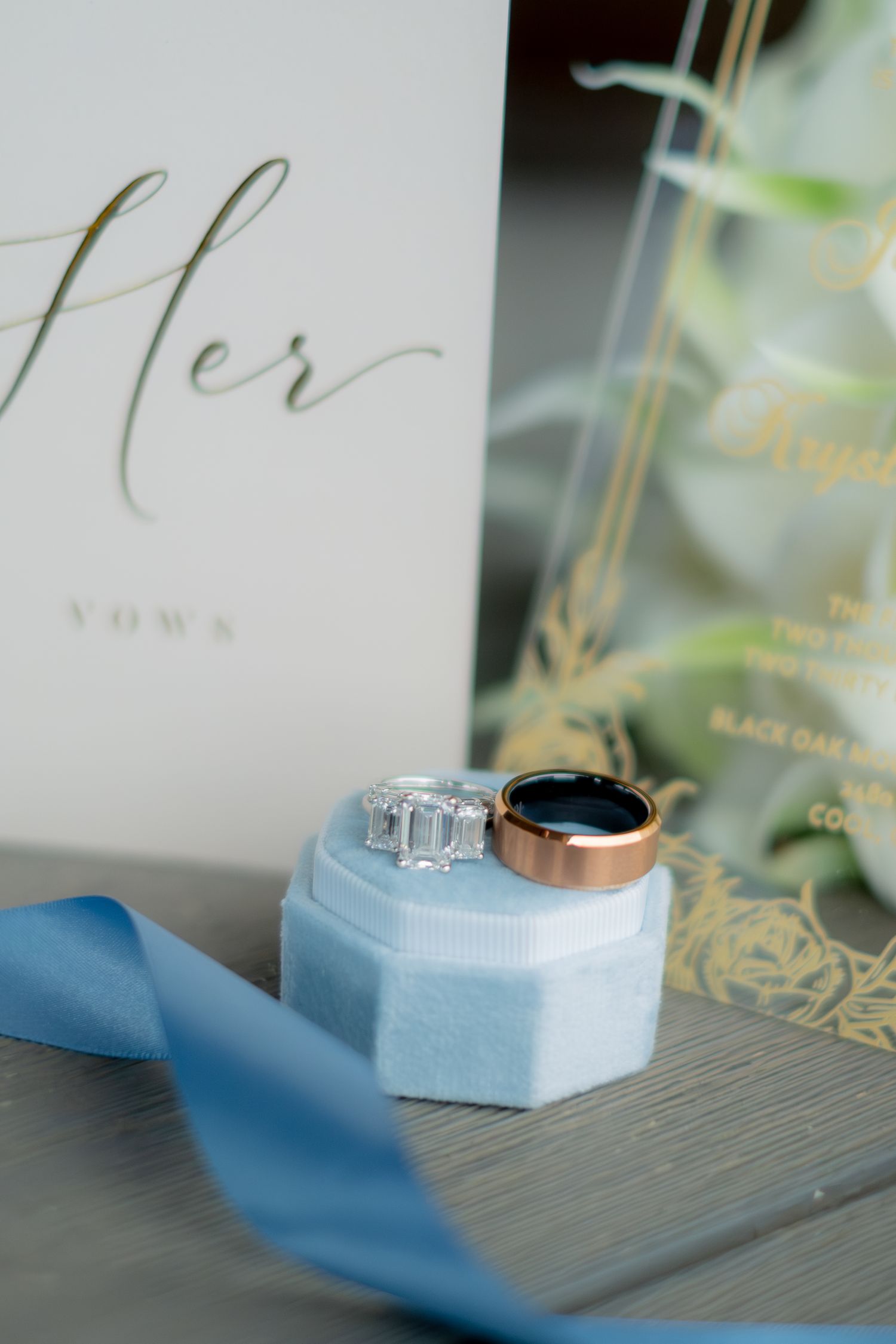 Wedding rings on a blue velvet box beside a vow book and gold floral invitation with blue ribbon.