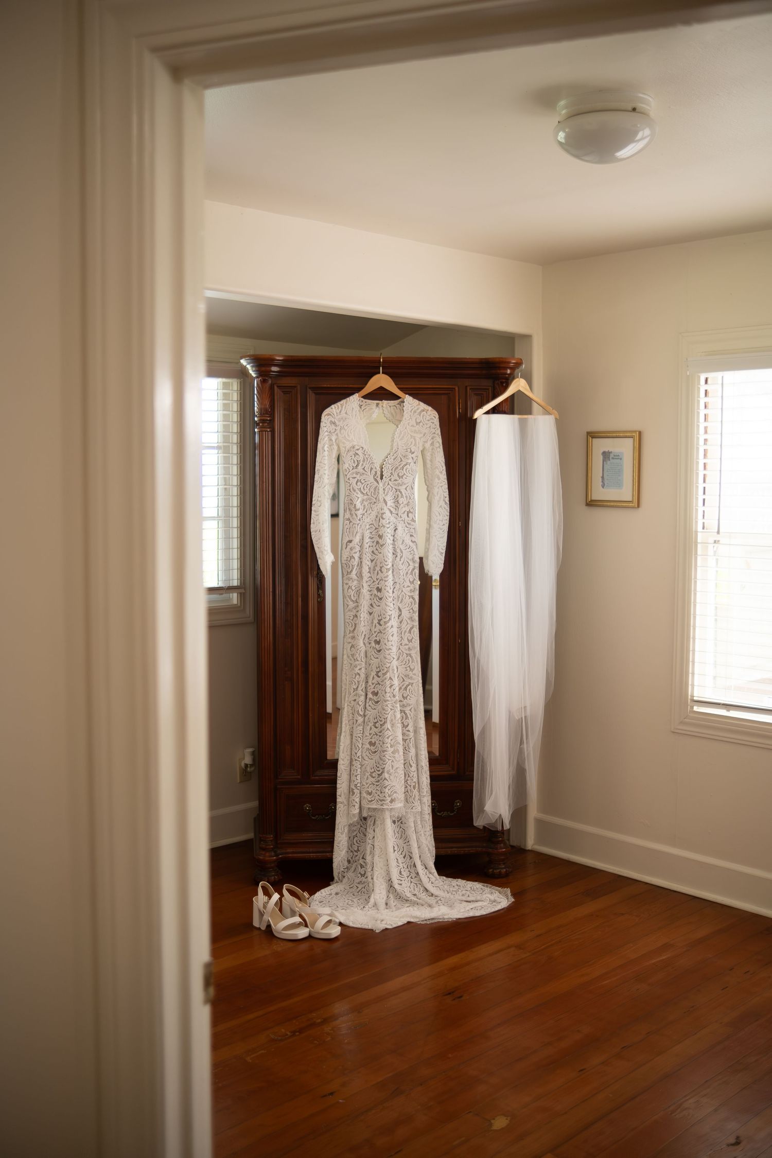 Elegant lace wedding dress and veil hanging on wooden armoire with bridal heels on hardwood floor.