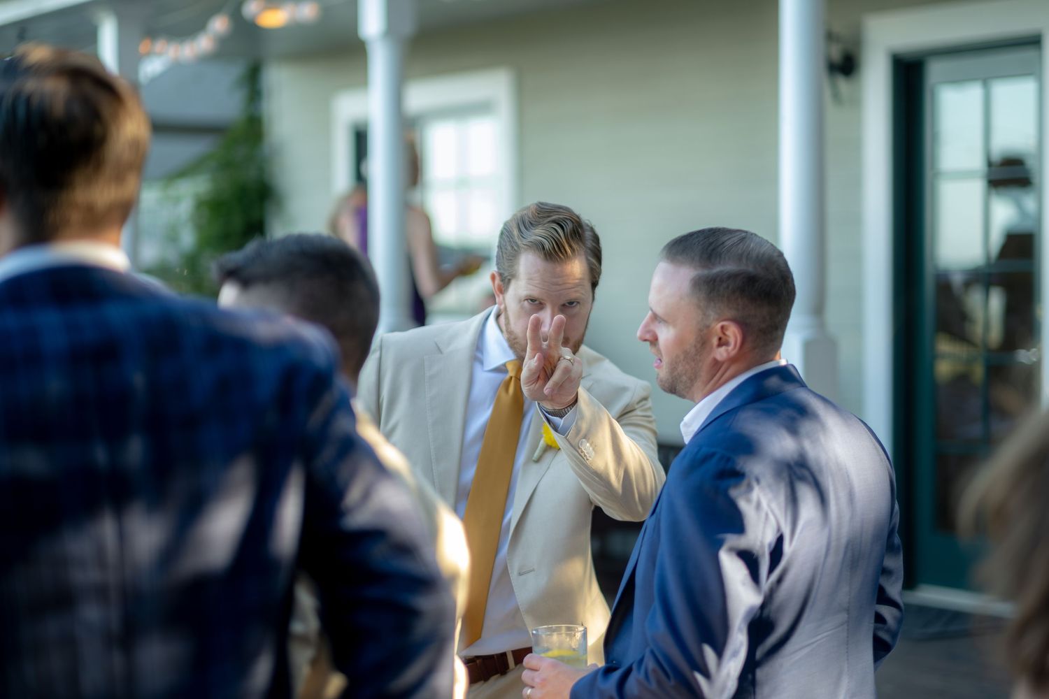 Groom in tan suit and yellow tie conversing with guests at an outdoor wedding reception.