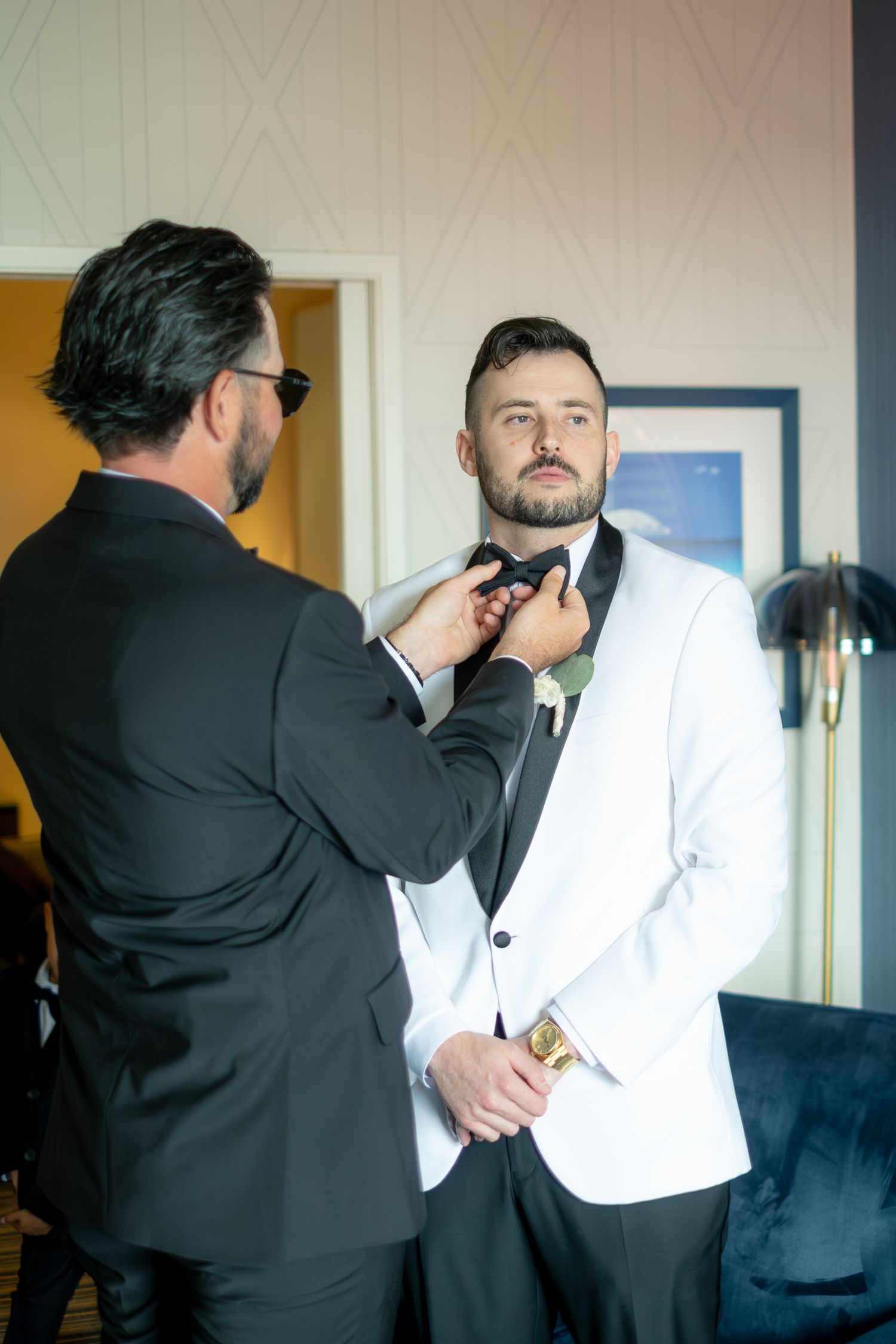 A groomsman adjusts the bow tie of a groom in a white tuxedo jacket before a wedding ceremony.