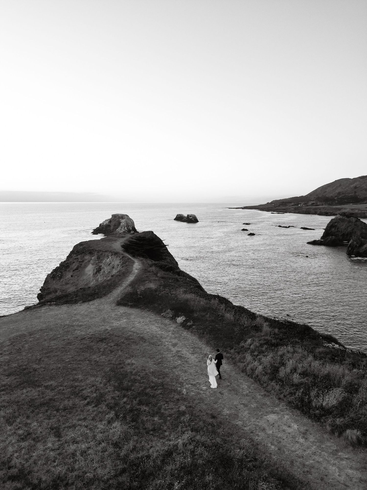 Aerial black and white photo of a wedding couple standing on a dramatic coastal cliff overlooking the ocean.