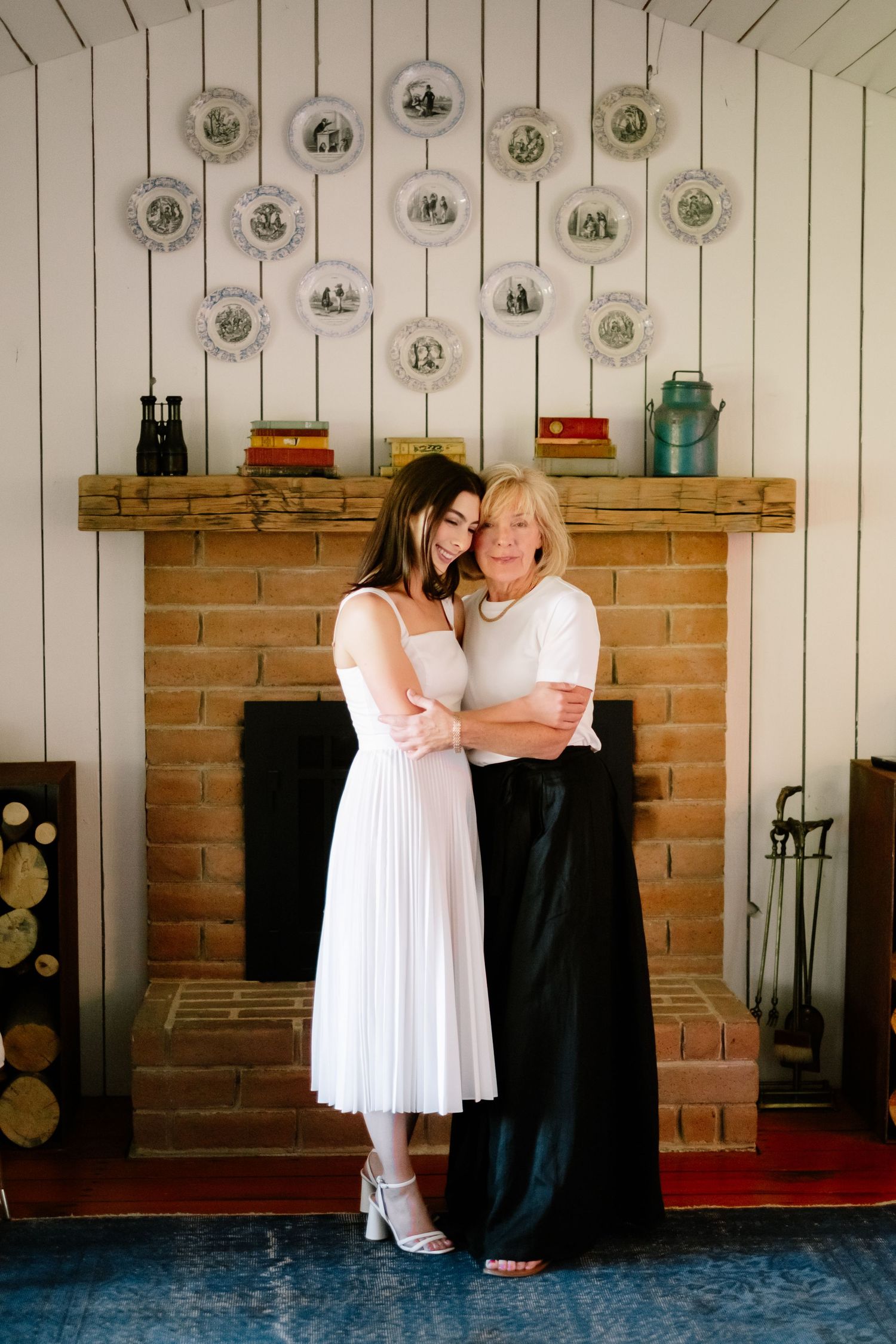 Bride in white midi dress embraces older woman near brick fireplace decorated with vintage plates.
