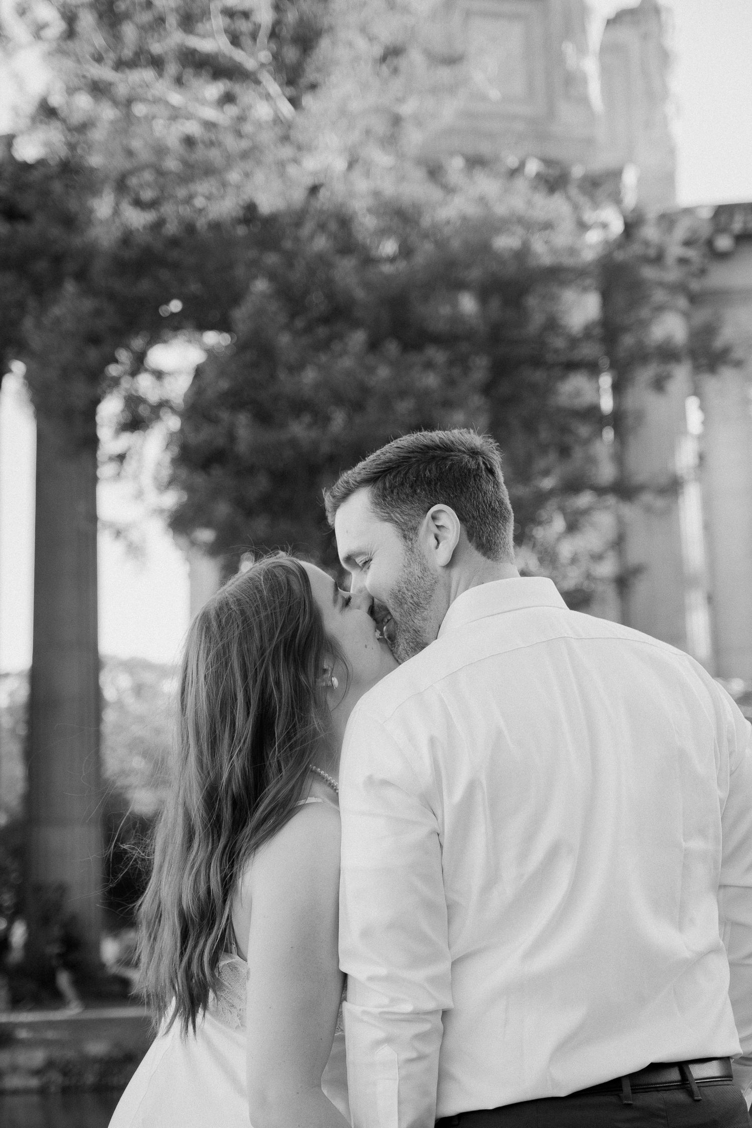 Black and white photo of a couple sharing a romantic kiss outdoors near elegant columns and lush trees.