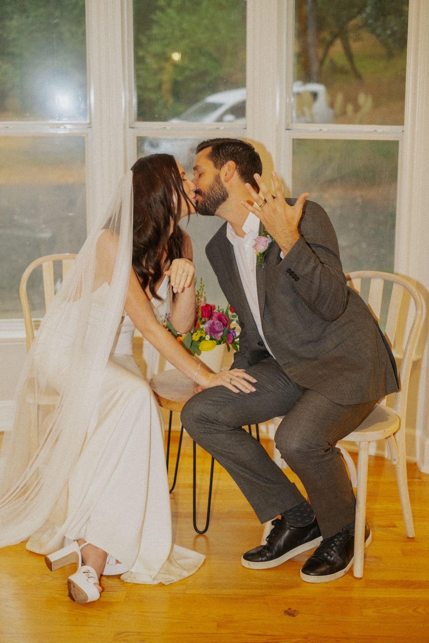 Bride and groom share a kiss while seated, showing off wedding rings, bride holding colorful bouquet in white gown and veil.