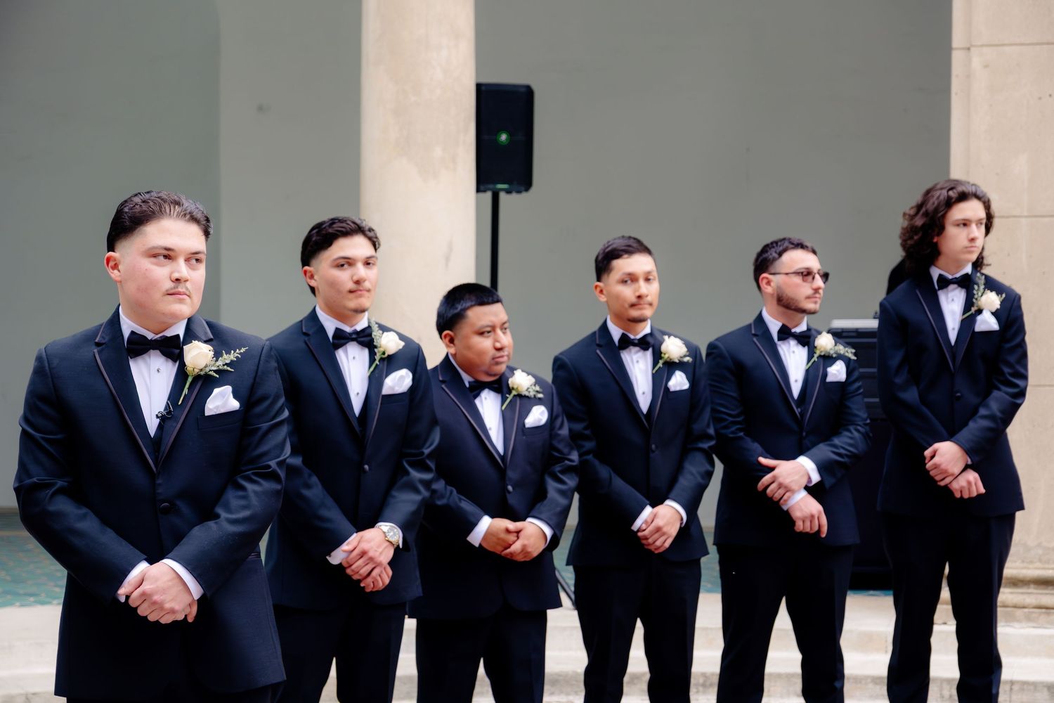 Six groomsmen in navy tuxedos with white boutonnieres and bow ties stand in a row at an elegant wedding venue.
