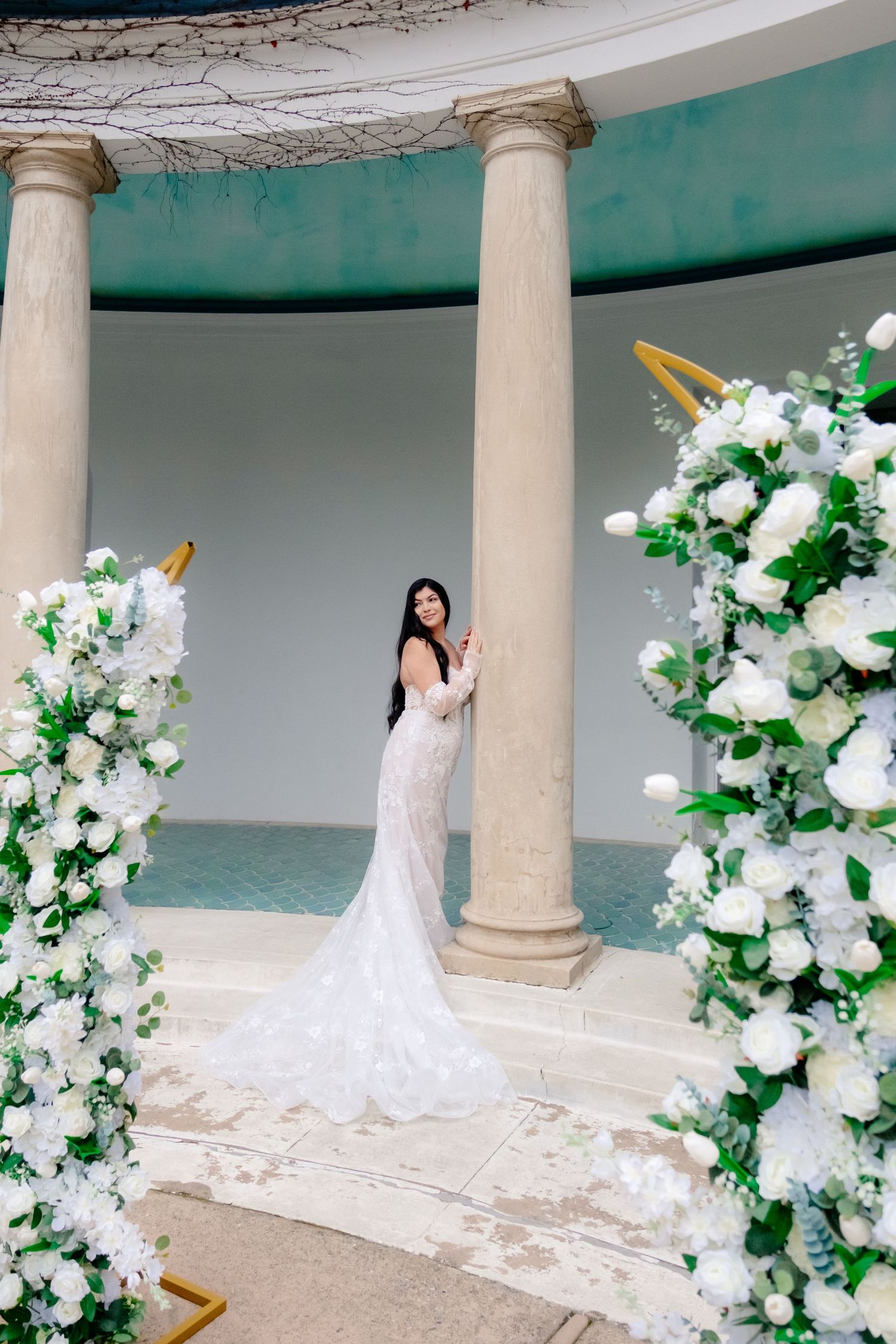 Bride in backless lace mermaid gown stands between white floral arrangements and stone columns.