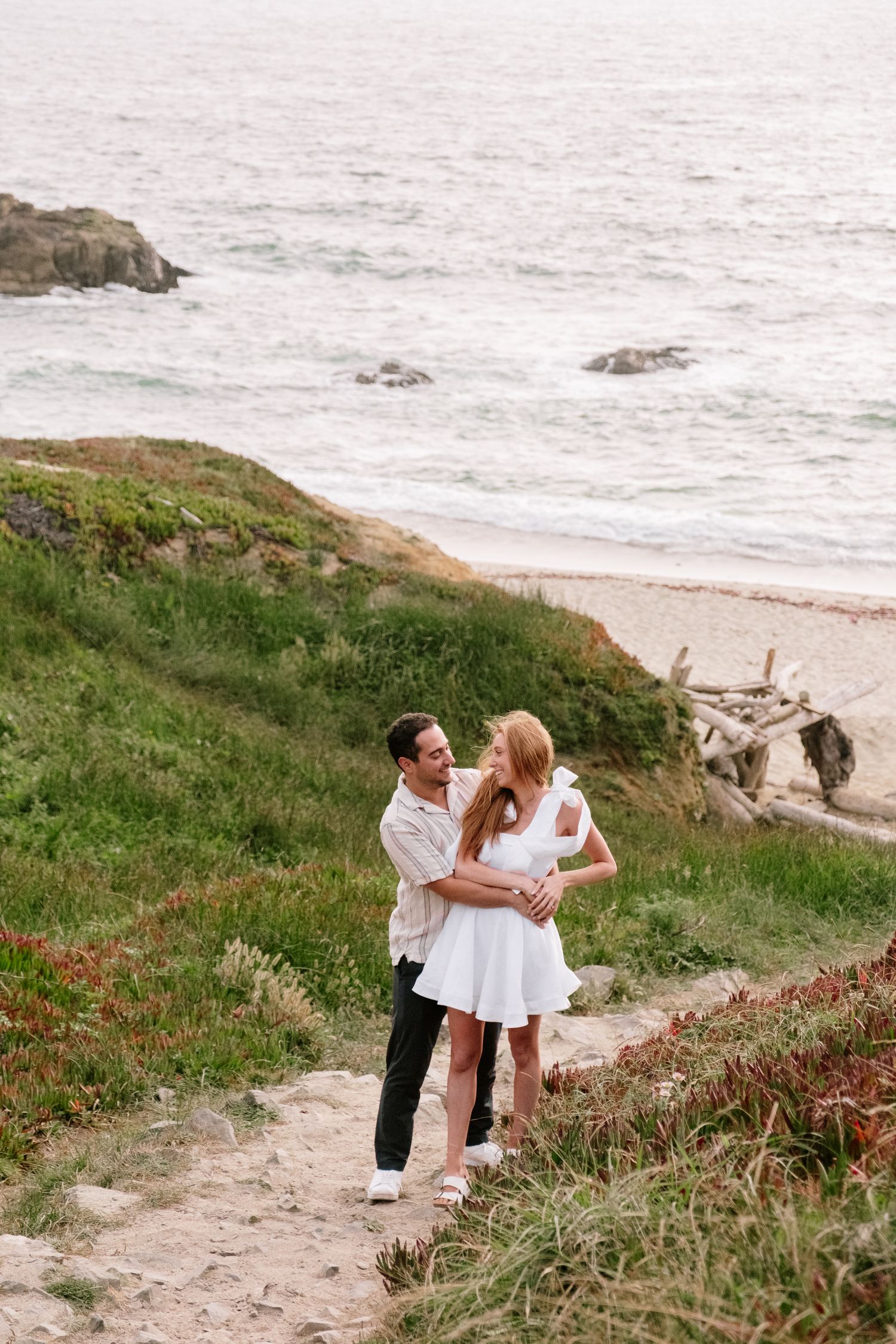 Romantic couple holding each other on a scenic coastal trail with ocean waves and sandy beach below.